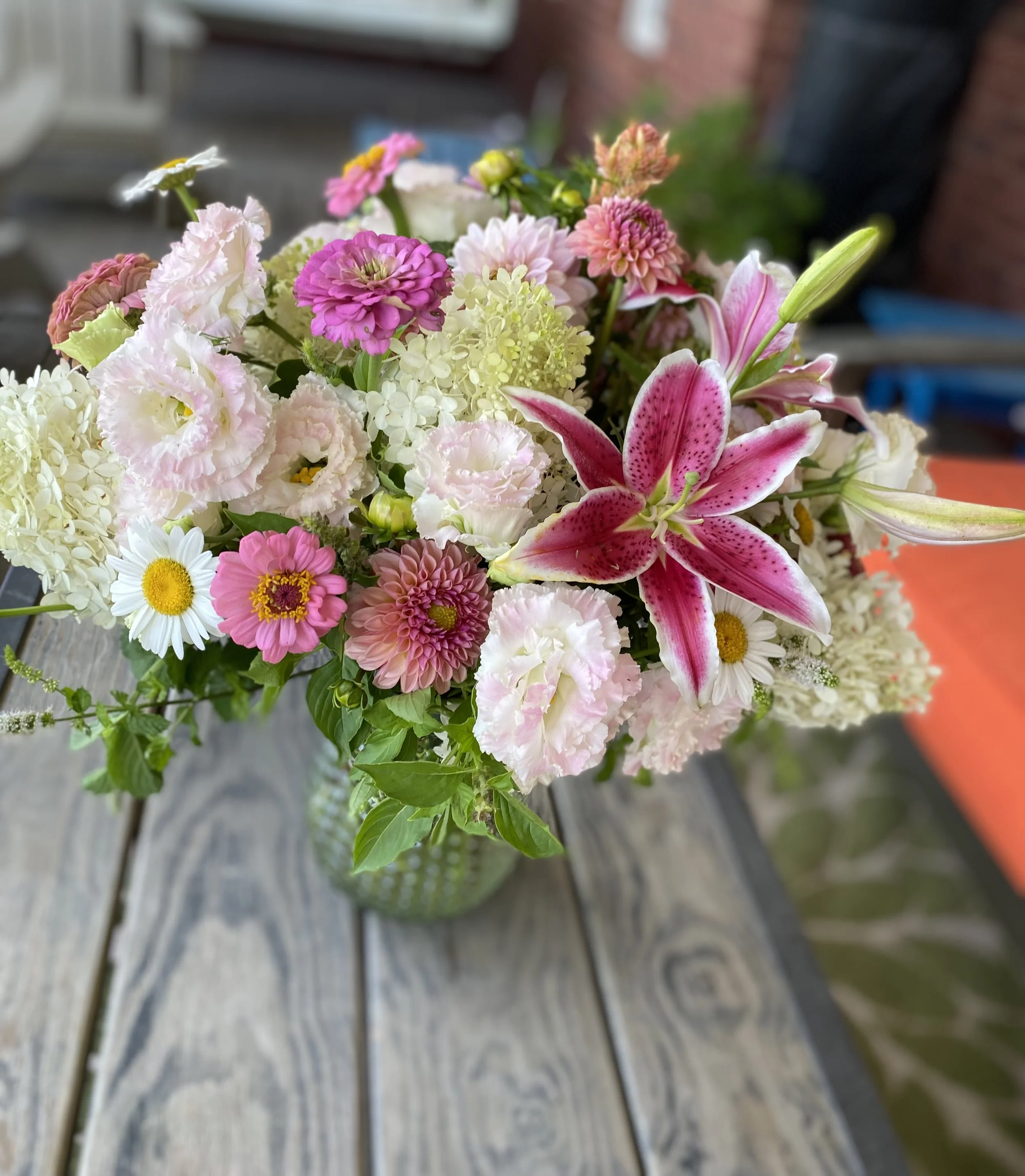 A colorful bouquet of various flowers including pink lilies, daisies, and hydrangeas in a glass vase on a wooden table.