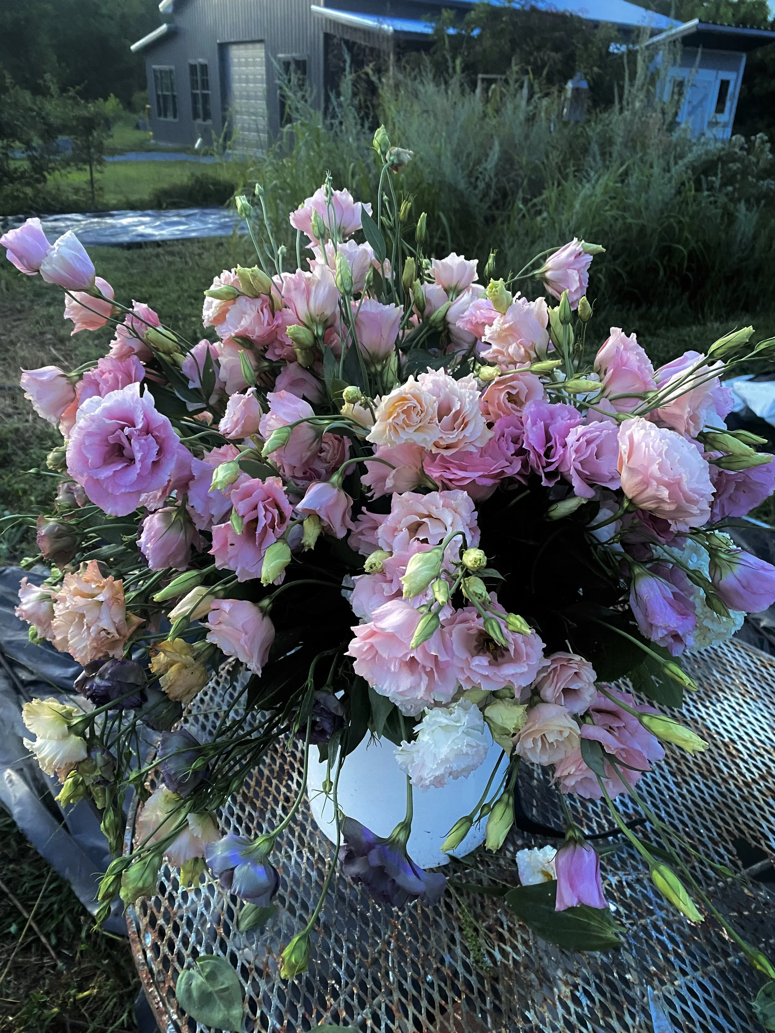 A bouquet of pink and purple flowers arranged in a white vase on a metal table outdoors, with a grassy yard and a building in the background.