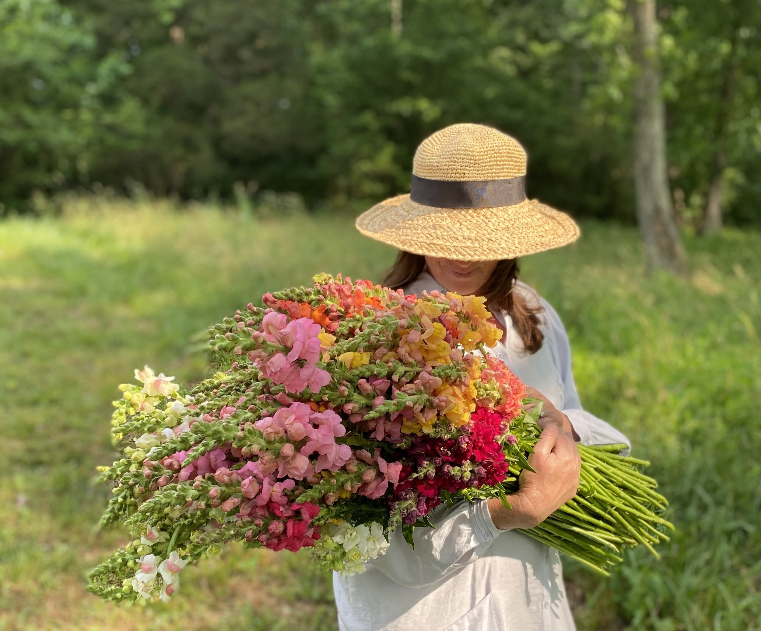 A woman wearing a large straw hat holding a large bouquet of colorful flowers outdoors in a green, wooded area.