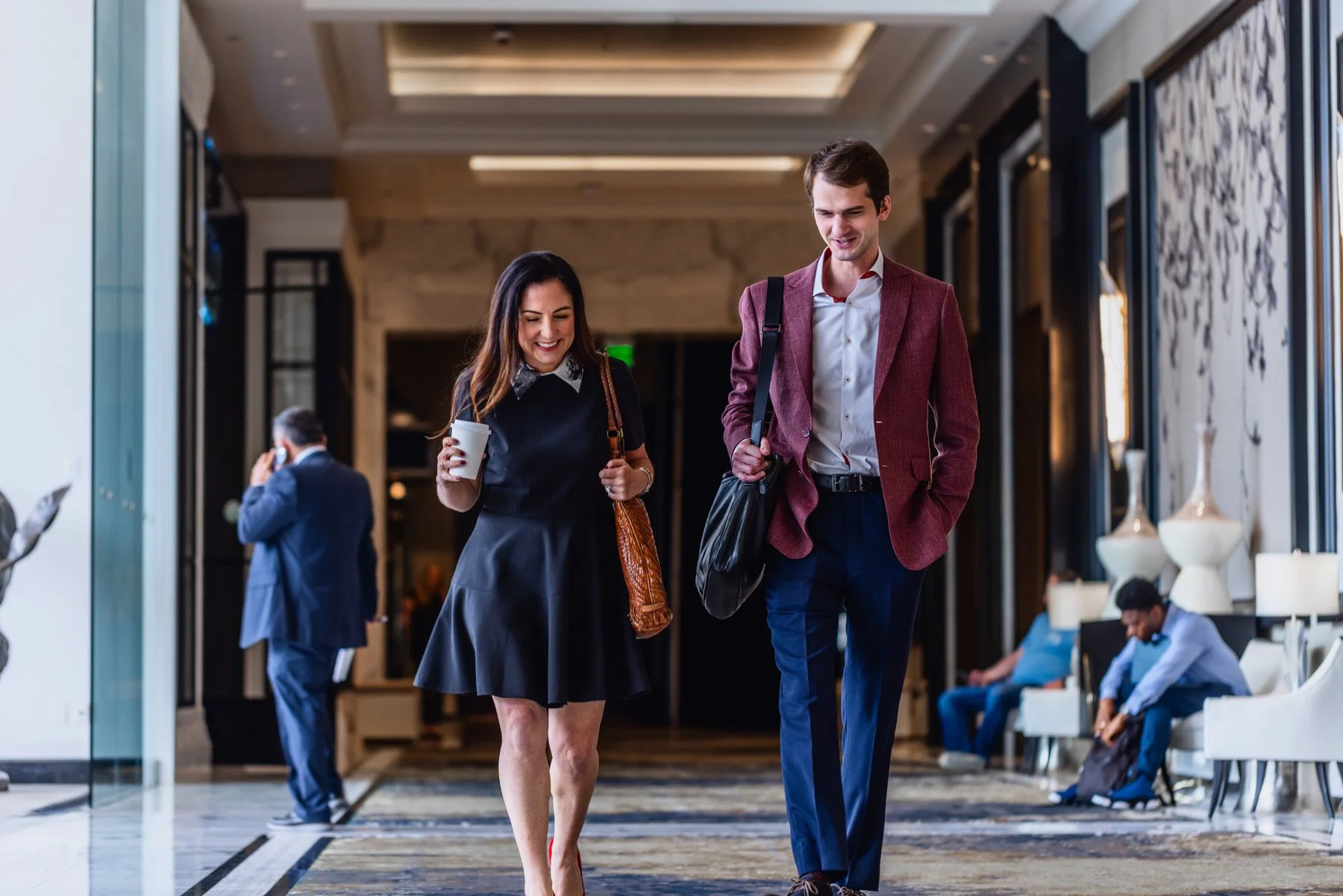 Two people walking and talking in a hotel lobby, a woman with a coffee cup and a man with a black bag, with other hotel guests in the background.