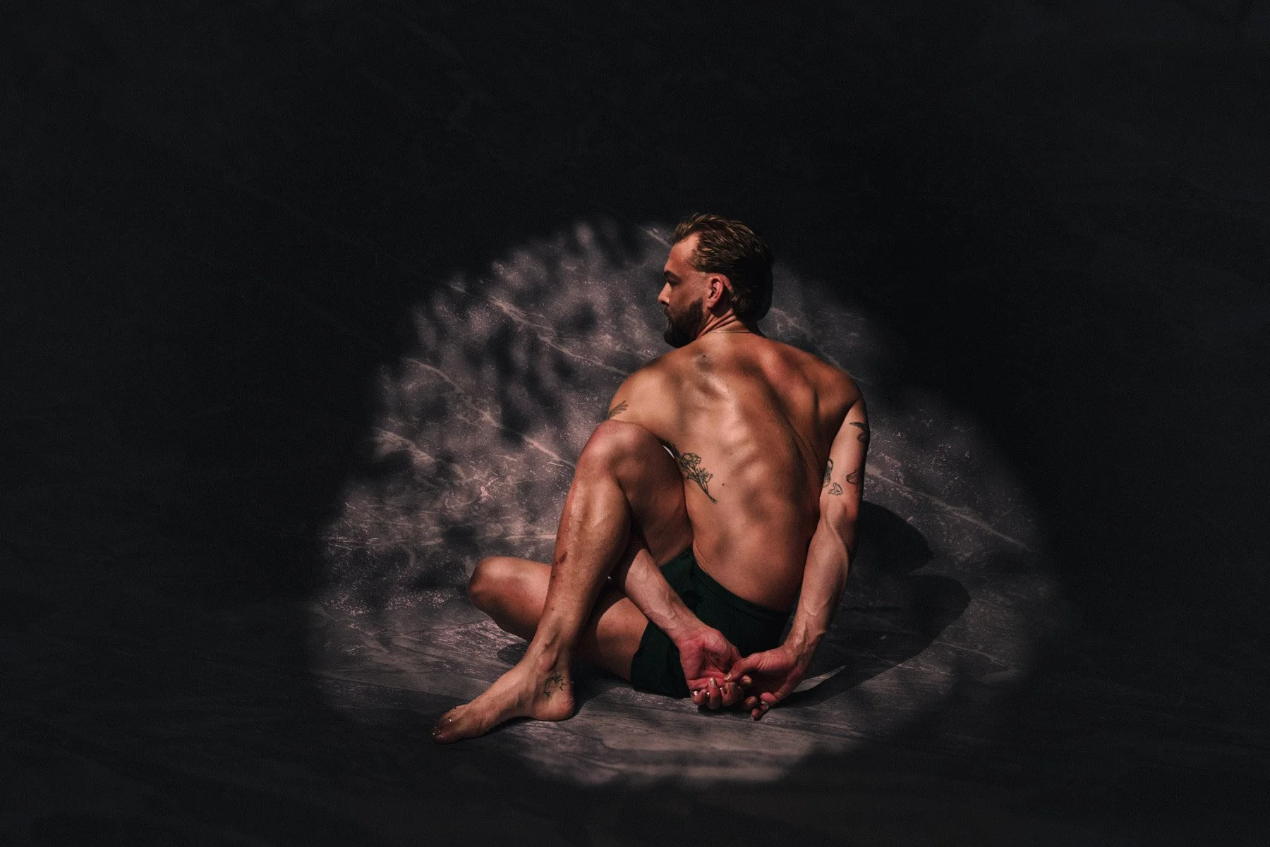 A shirtless man with tattoos sitting in a yoga pose against a dark background with pink powder or dust. The man appears to be balancing with one arm behind his back, holding his foot with both hands.