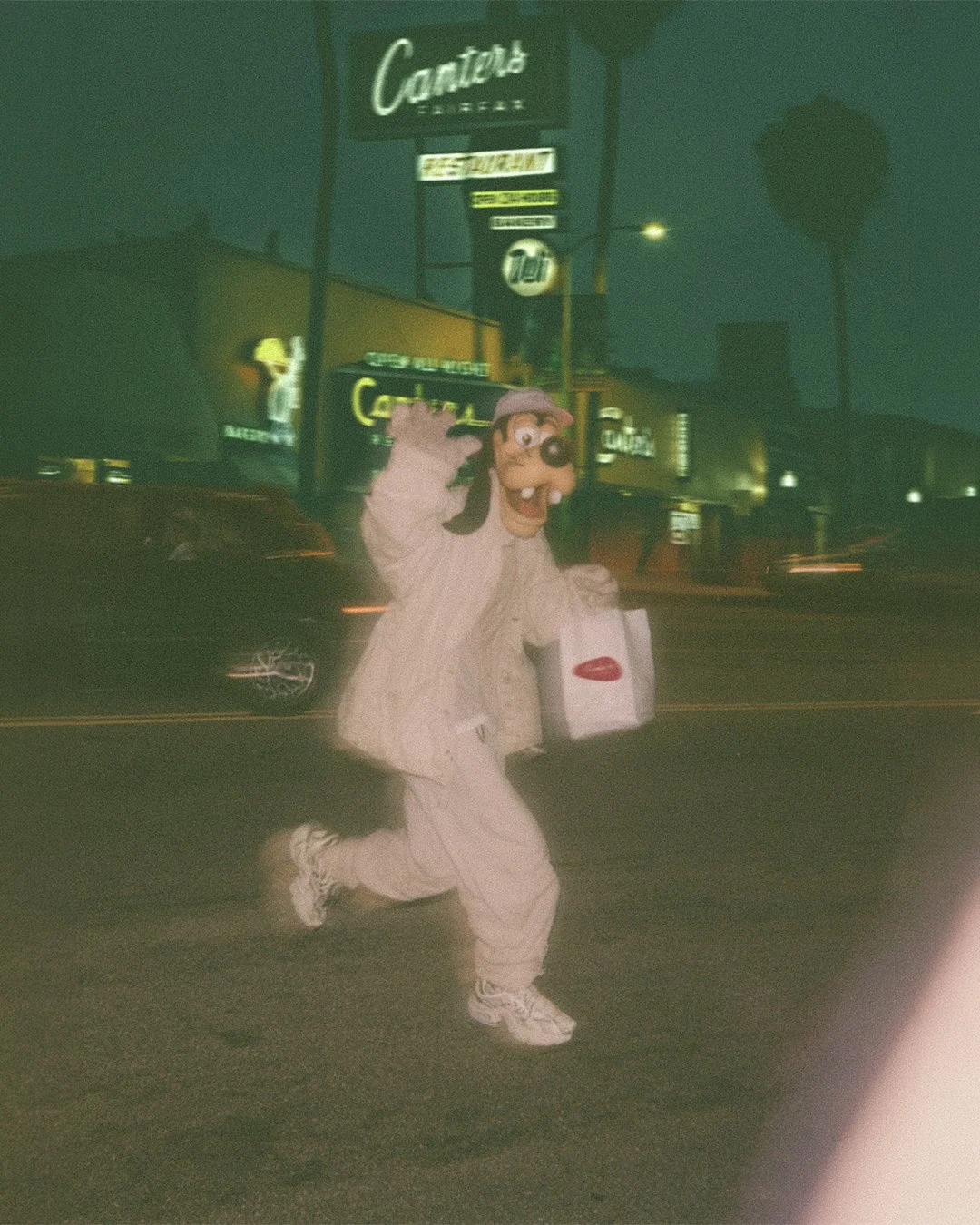 A person dressed in a Goofy costume from Disney is running across a street at night, carrying a Canters bag