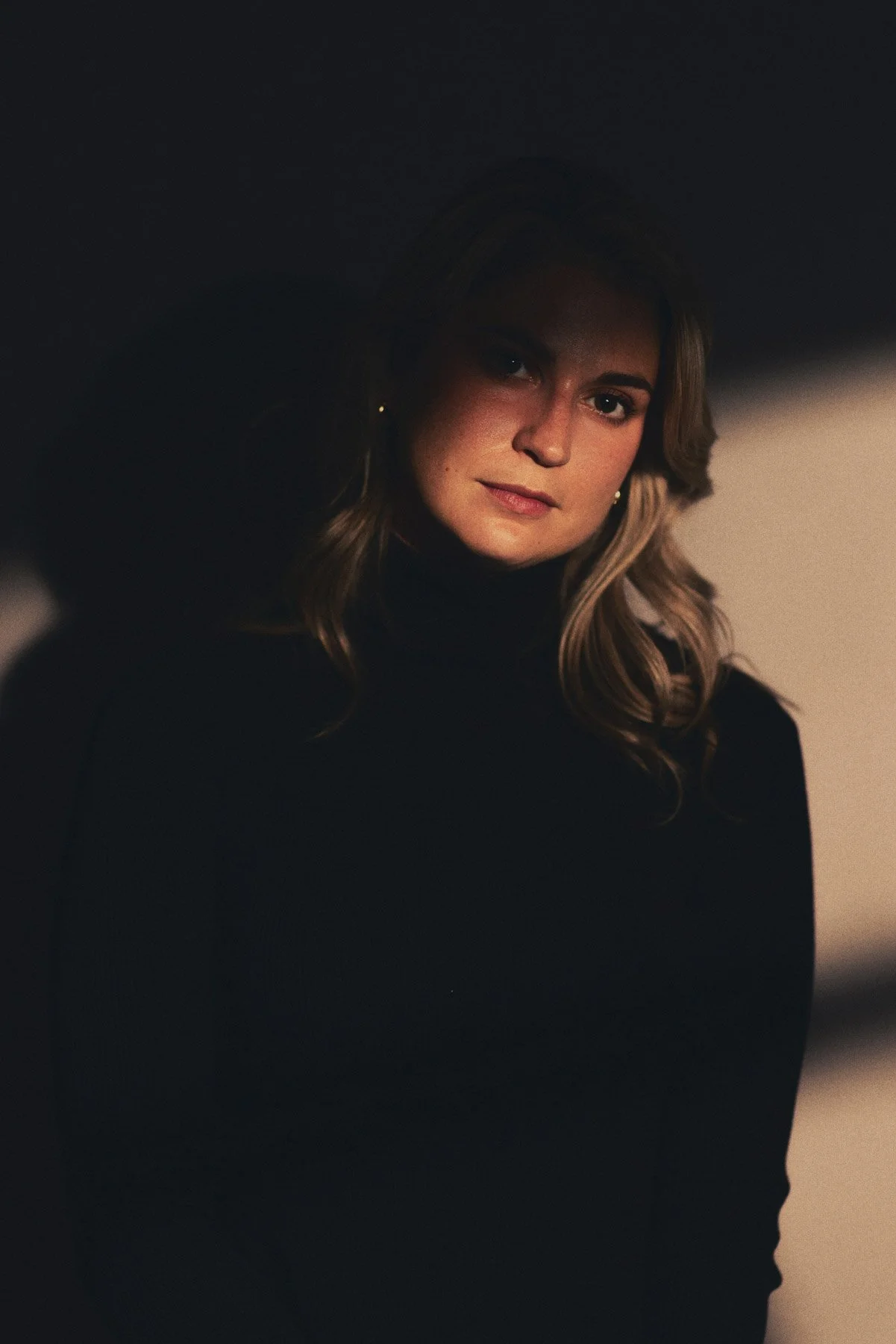 A woman with wavy light brown hair and earrings, wearing a black turtleneck, looking at the camera with a neutral expression in a dimly lit setting.
