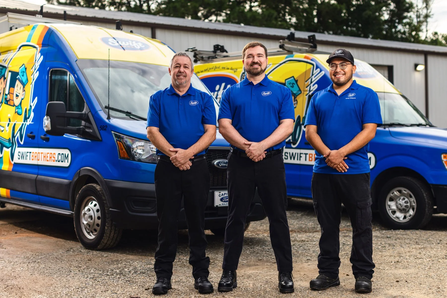 Three men standing in front of two blue Swift Brothers service vehicles, all wearing matching blue polo shirts with the Swift logo, smiling for the camera in an outdoor setting.
