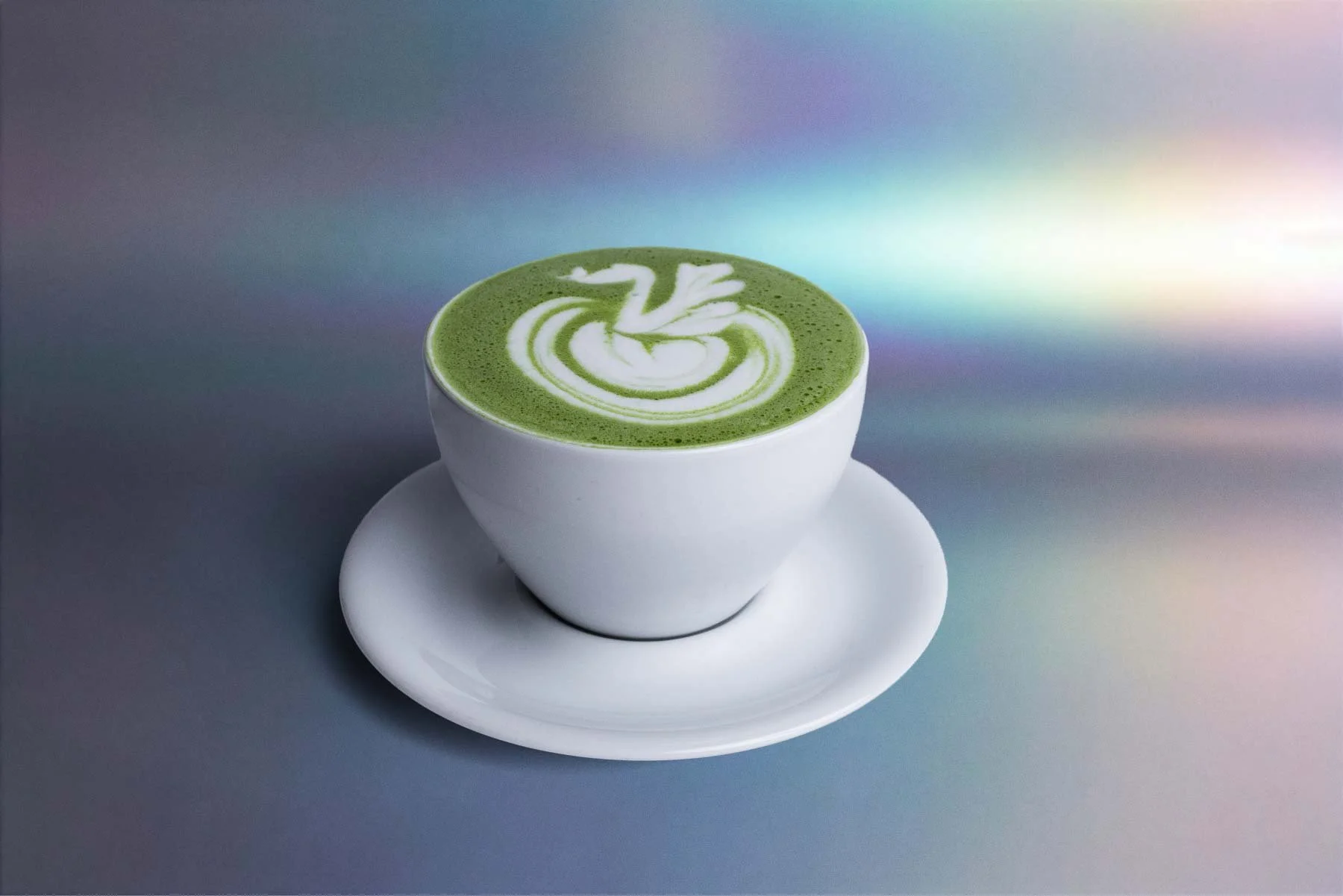 A white cup filled with green matcha latte, featuring intricate white latte art on top in the shape of a swan, with a matching white saucer underneath. The background displays a gradient with holographic colors.