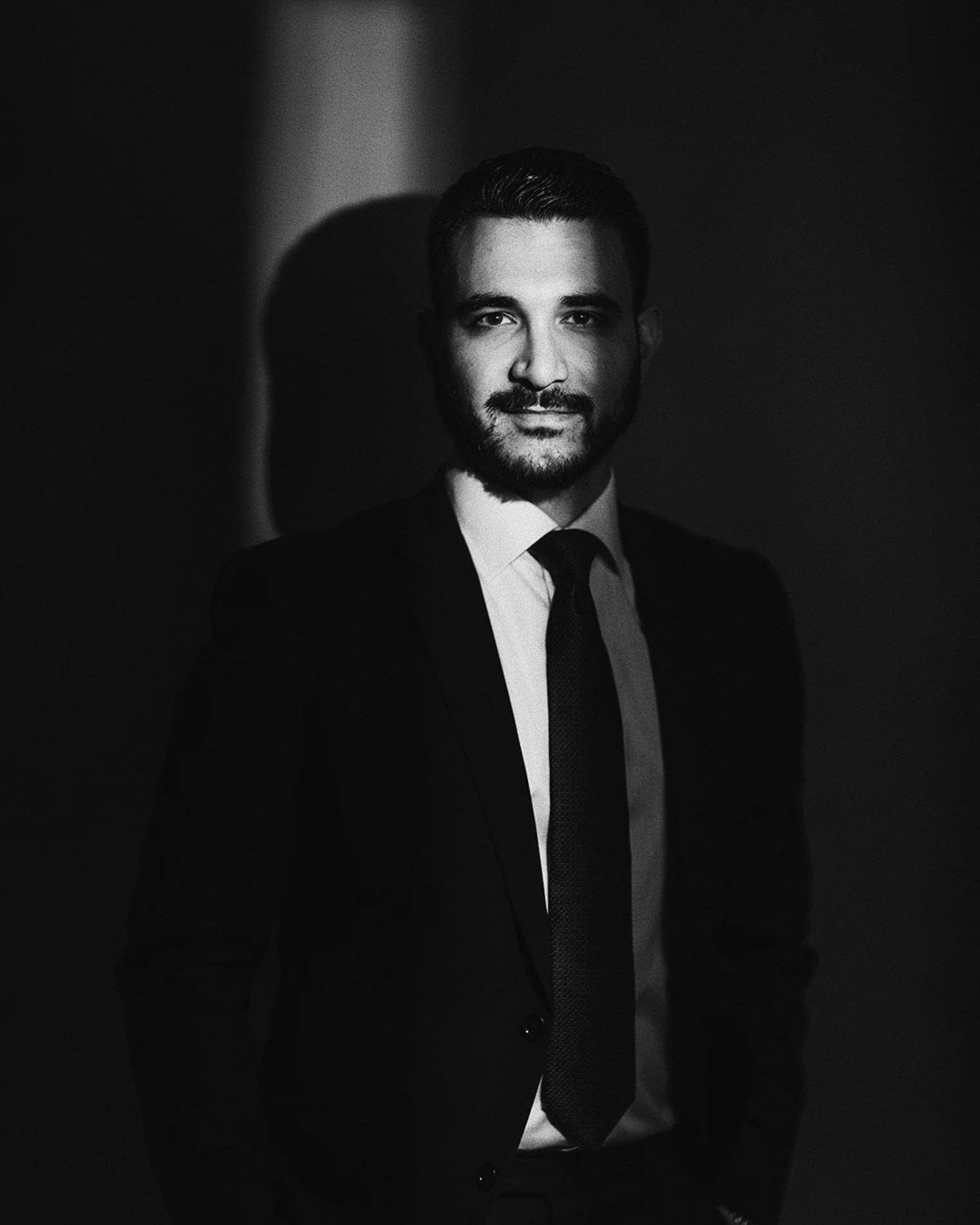 Black and white portrait of a man wearing a suit and tie, smiling slightly, with a shadow cast on the dark background.