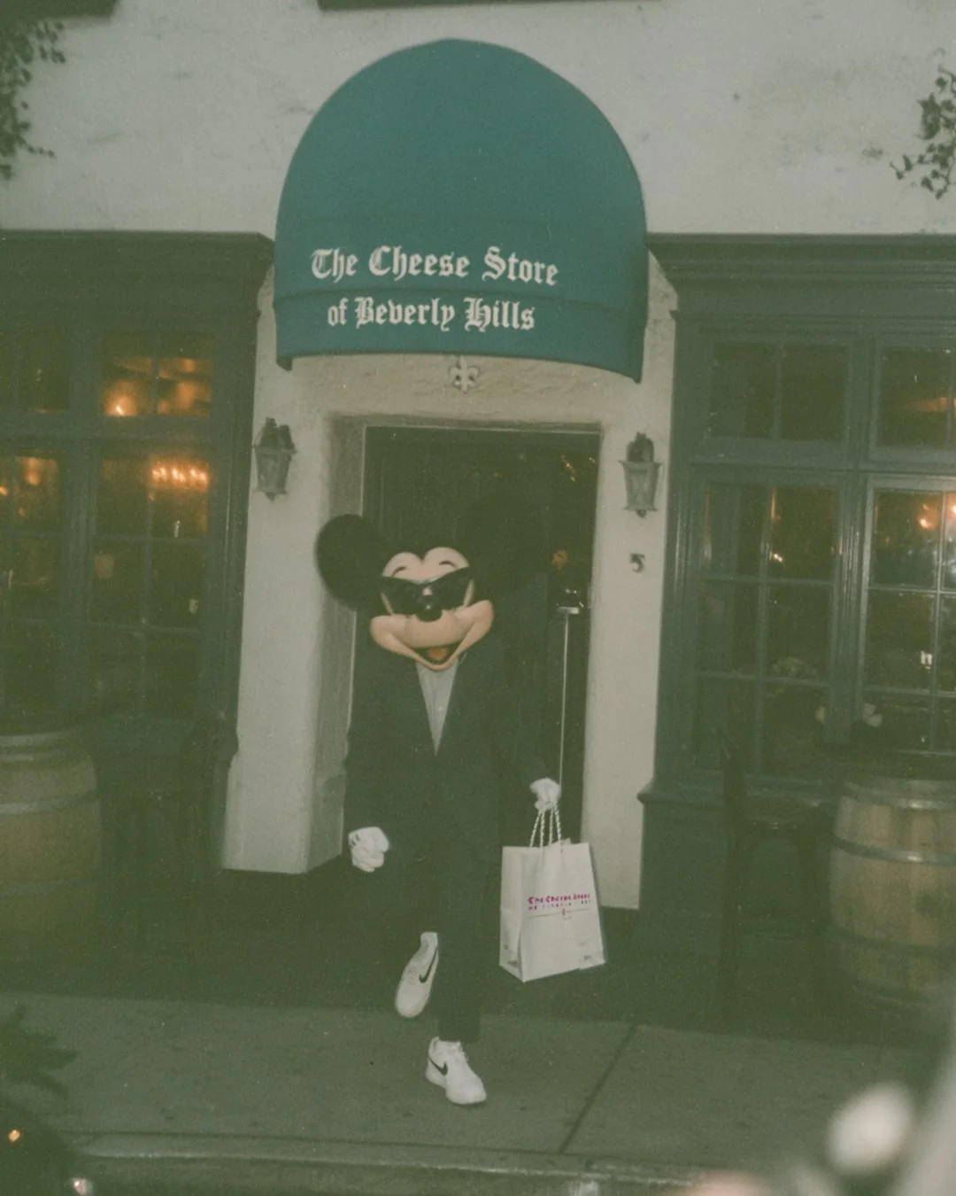 A person dressed as Mickey Mouse wearing sunglasses, a black suit, and white sneakers, standing outside The Cheese Store of Beverly Hills at night, holding a shopping bag, in front of a store entrance with a green awning.