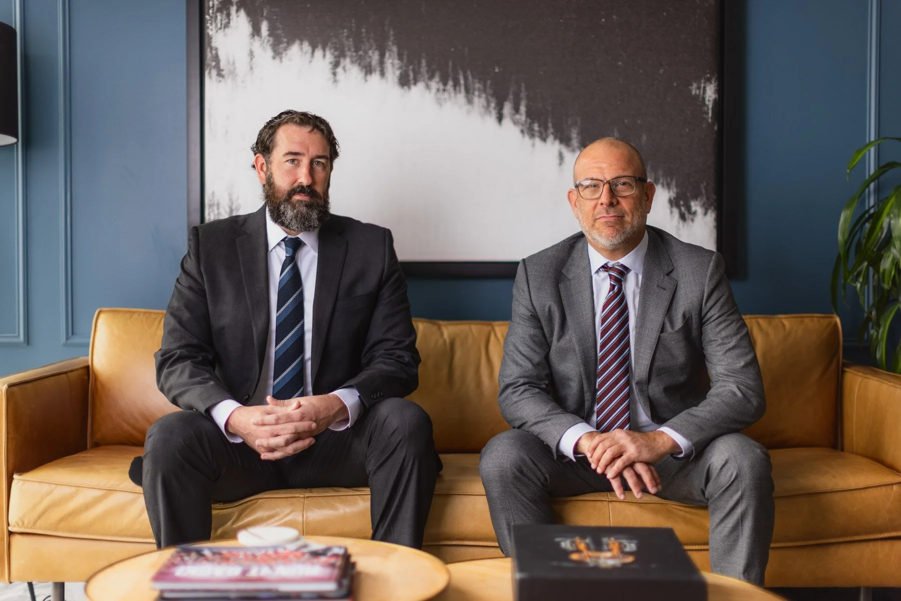 Two men in suits sitting on a yellow leather sofa in a modern office or lobby, with a large abstract black-and-white painting behind them. The man on the left has a beard and dark hair, while the man on the right is bald and wearing glasses.