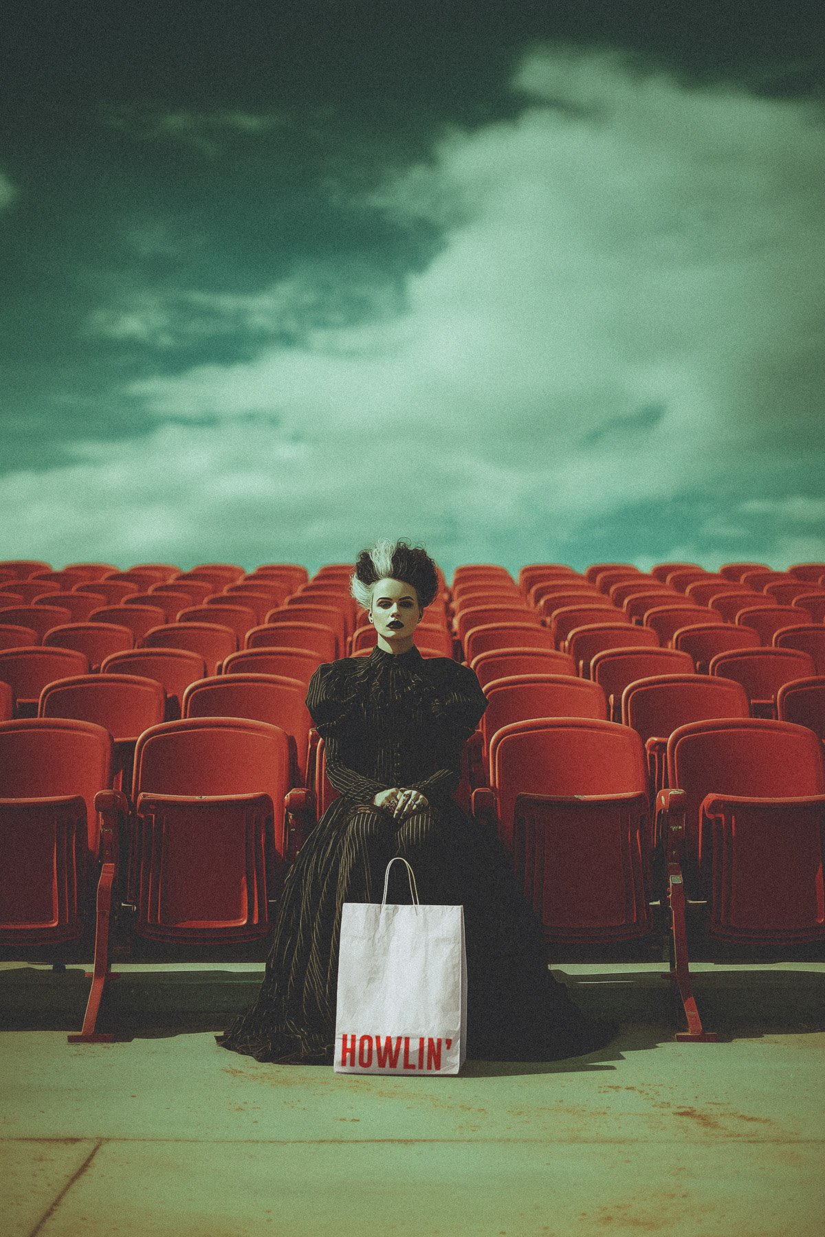 A person with dark and white curly hair and dark makeup sitting in a nearly empty outdoor theater or stadium with red seats, under a cloudy sky. They are wearing a dark Victorian-style dress with puffed sleeves, and there is a shopping bag labeled "H