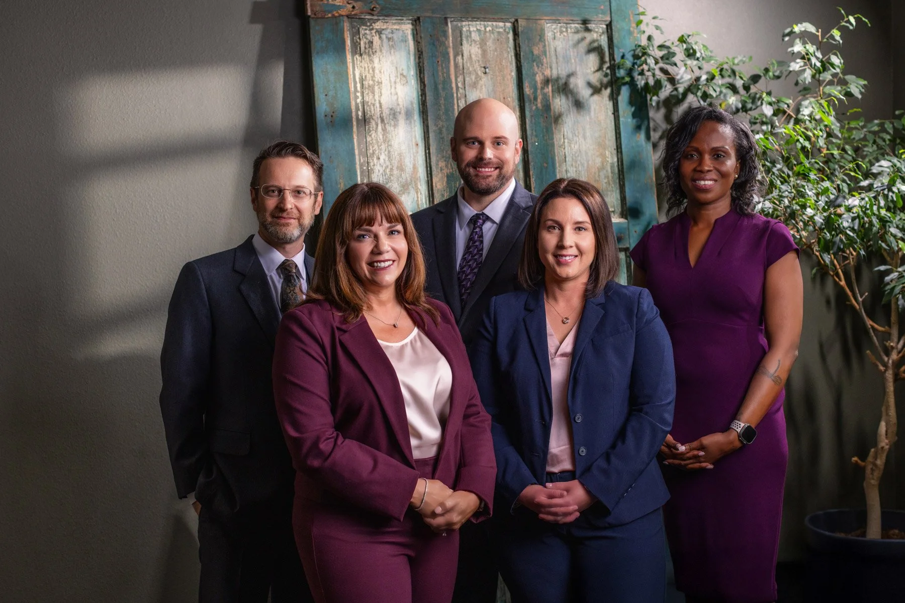 A diverse group of five professionals, three women and two men, dressed in business attire, posing together indoors in front of a rustic background with green plants.