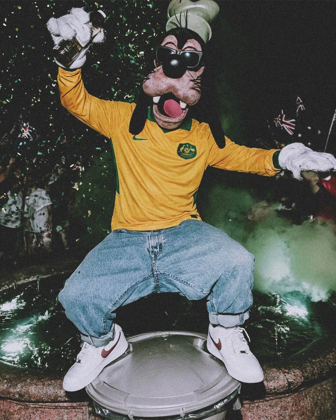 Goofy with sunglasses, yellow Australia jersey, baggy jeans, and white sneakers, standing on a drum and striking a lively pose in front of green fireworks and lights.
