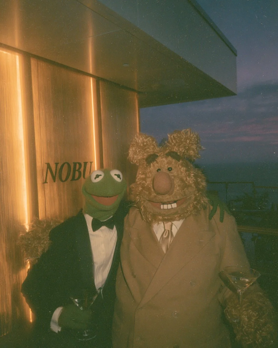 Kermit the frog and a bear, dressed in formal tuxedos with bow ties, standing outside at night, holding drinks in martini glasses. The background shows a wooden wall with the word 'NOBU' and a cityscape with a darkening sky.