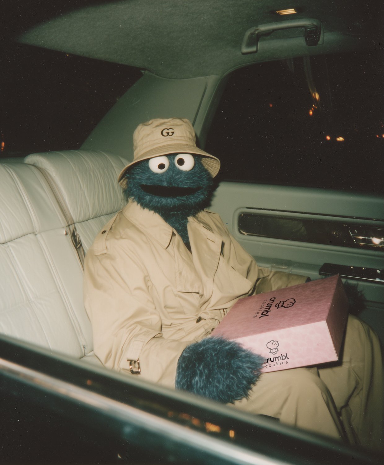 Cookie Monster from The Muppets dressed as a detective, sitting in a car at night, holding a box of crumble cookies.