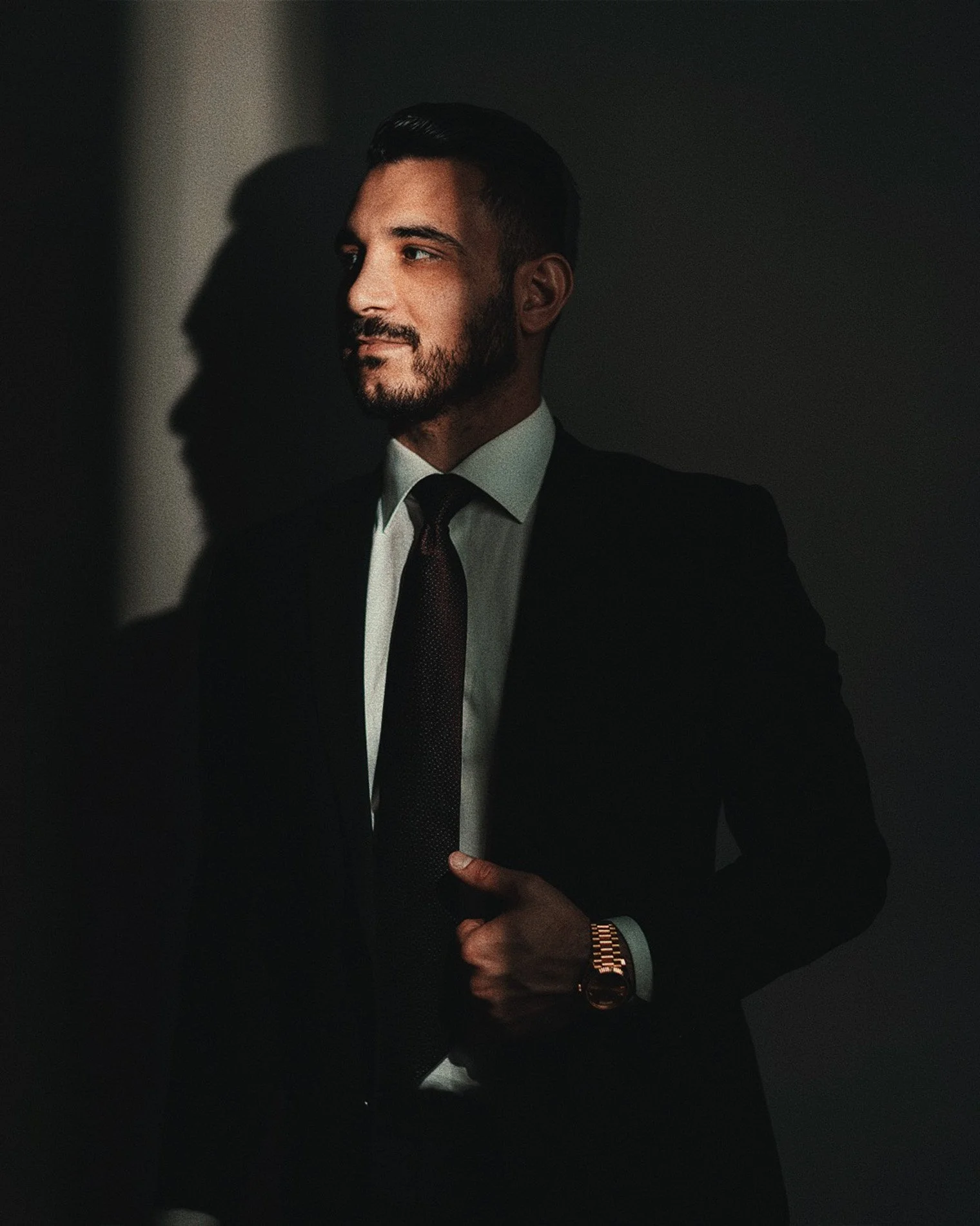 A man in a suit and tie with a beard and slicked-back hair standing in a dark room with a shadow of his profile cast on the wall, illuminated by soft lighting.