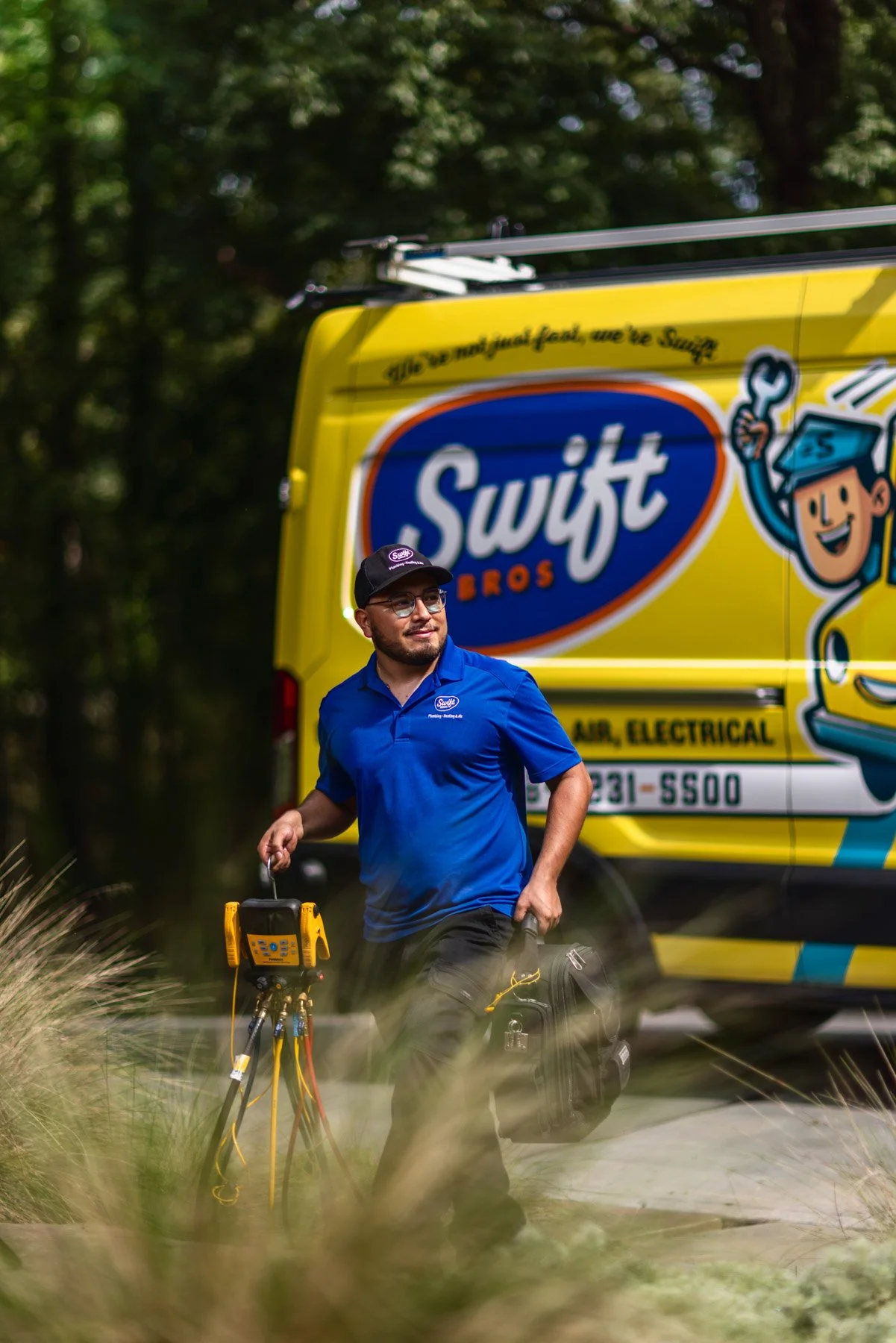 A technician wearing a blue uniform and cap, holding electrical testing equipment, standing outdoors in front of a bright yellow Swift Bros service van with cartoon graphics, surrounded by greenery.