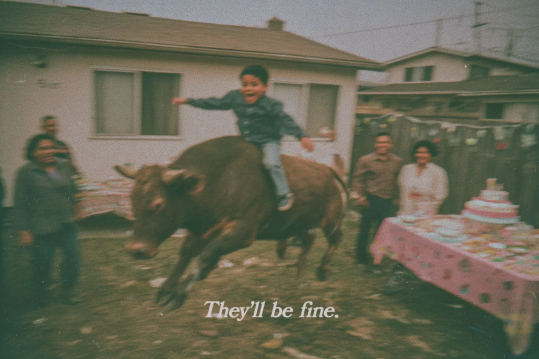 A young boy rides a bucking bull at a backyard celebration with a birthday cake on a table, surrounded by adults, against a residential house background.
