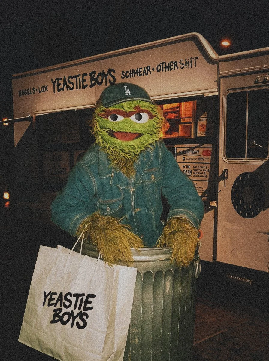 A Yeastie Boys street food truck at night with Oscar the Grouch in a trashcan, holding a shopping bag labeled 'Yeastie Boys'. the food truck has a sign with various food items listed.