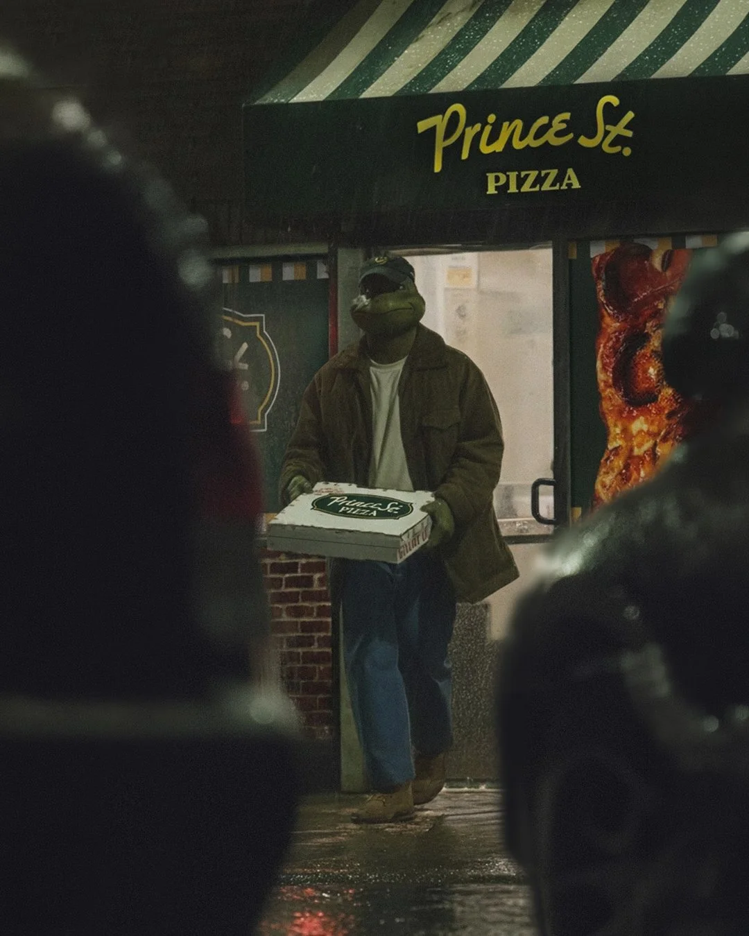 Ninja Turtle Leonardo holding a pizza box outside a pizza shop called Prince St. Pizza at night.