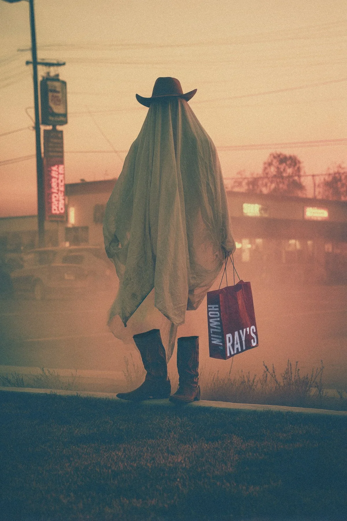 A person dressed as a cowboy, wearing a hat and long coat, holding shopping bags labeled "Ray's" and "Hometown," standing on the sidewalk during sunset or dusk.
