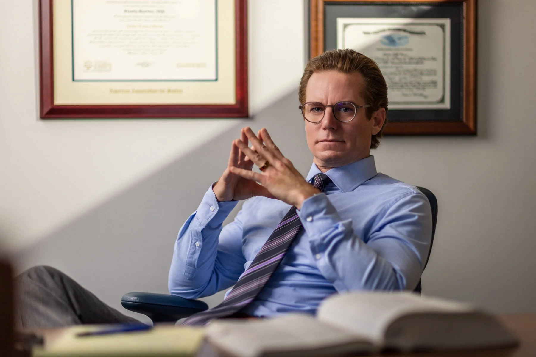 A man with glasses wearing a blue shirt and a striped tie sitting in an office, with framed certificates on the wall behind him.