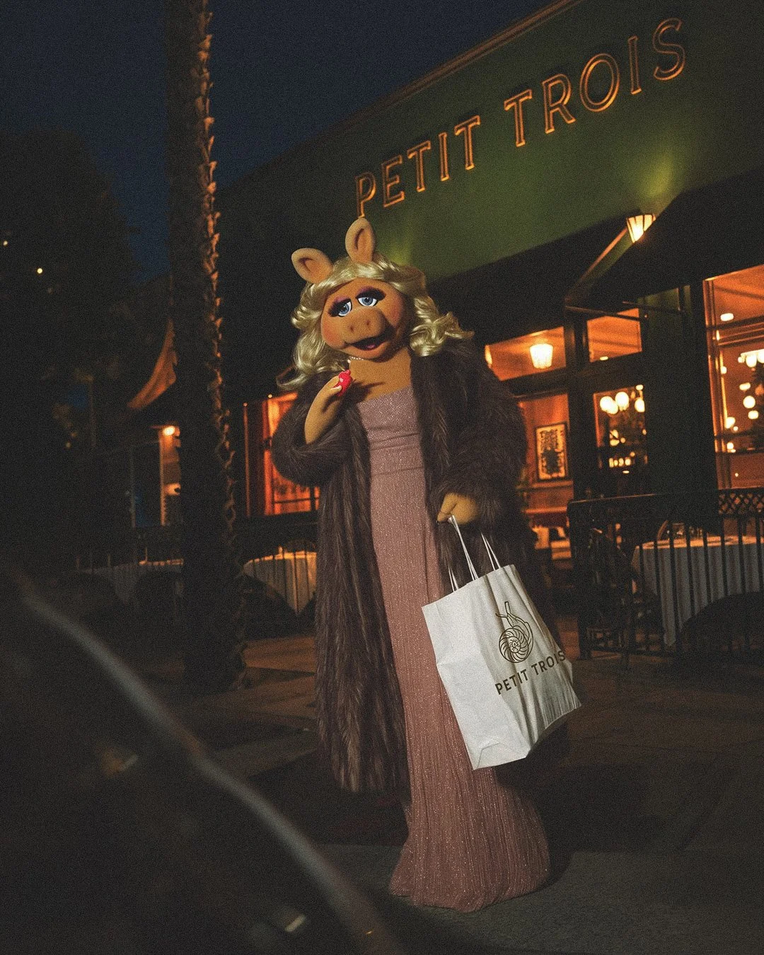 Miss Piggy standing outside a restaurant called 'Petit Trois' at night, holding a shopping bag from 'Petit Trois'.