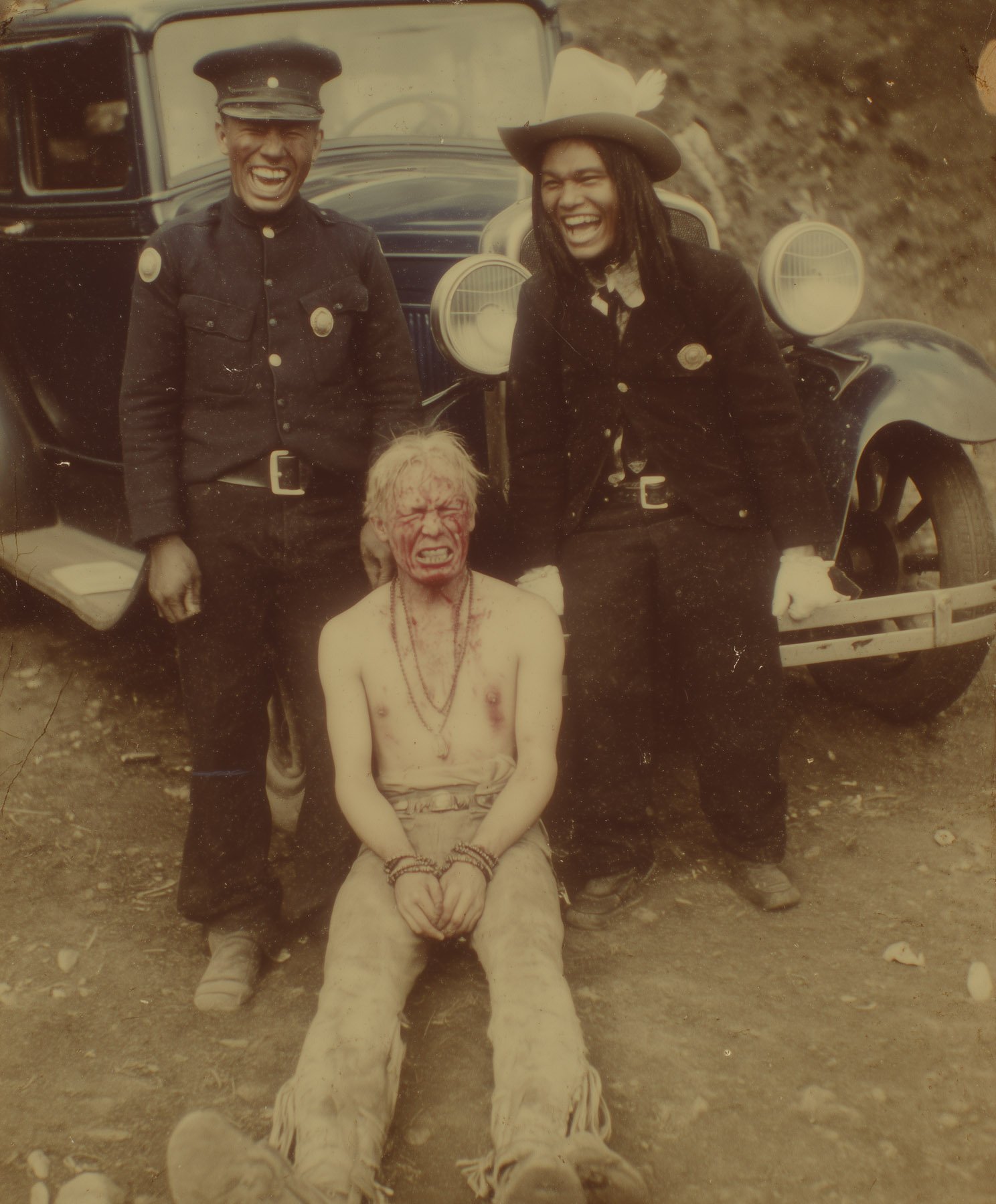 Black and white photo of two police officers standing in front of a vintage car, laughing, with a shirtless man sitting on the ground in front of them, covering his face with his hands and grimacing, with dirt and rocks on the ground.