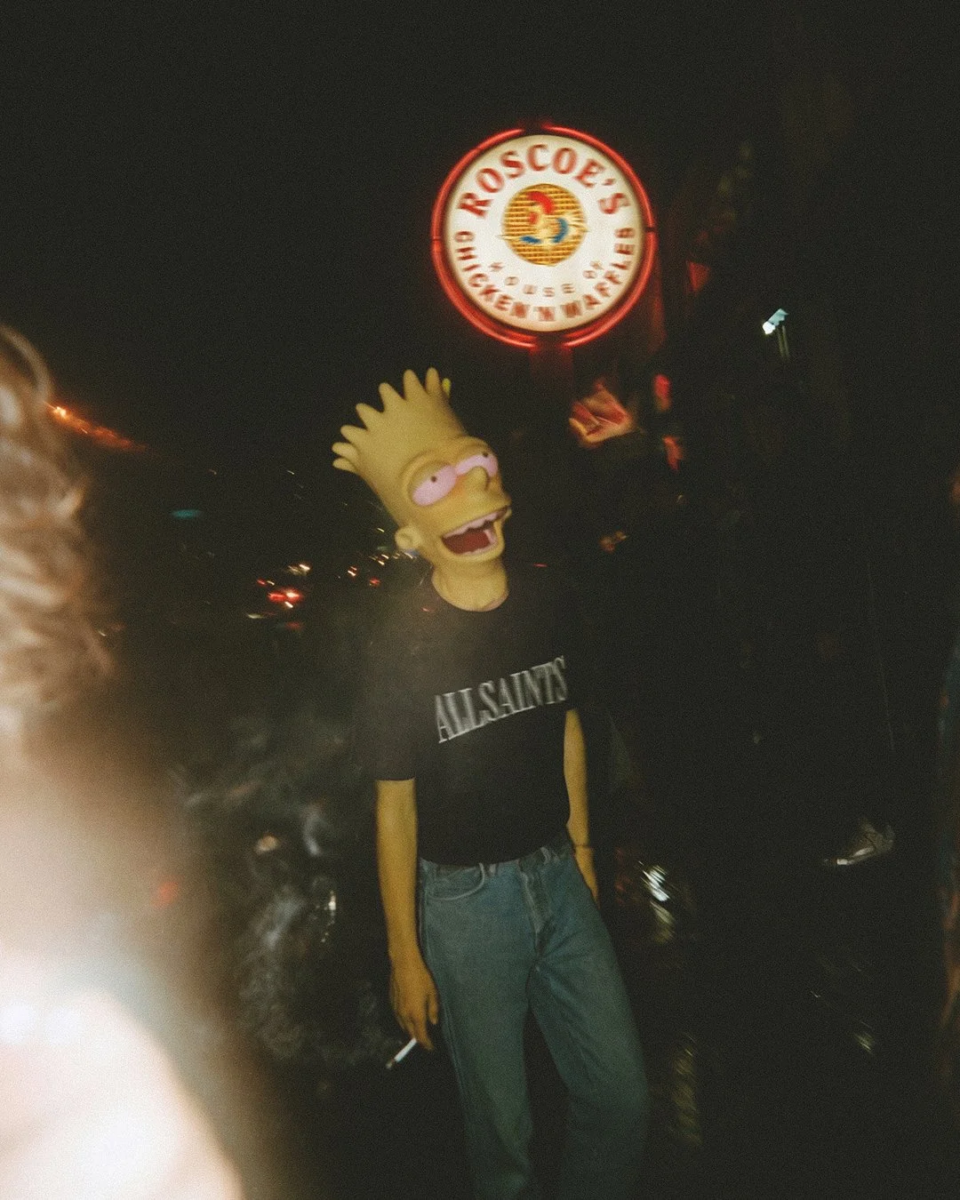 Bart Simpson standing at night near a neon sign for Roscoes Chicken and Waffles. The person is dressed in a black t-shirt with white text and blue jeans, holding a cigarette in one hand.