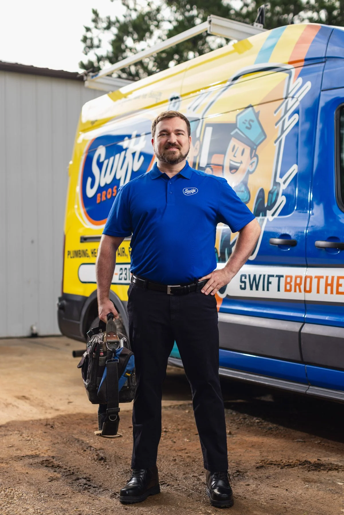 A man in a blue uniform standing in front of a blue and yellow Swift Brothers utility van, holding a tool bag, outdoors on a dirt surface with a building and trees in the background.