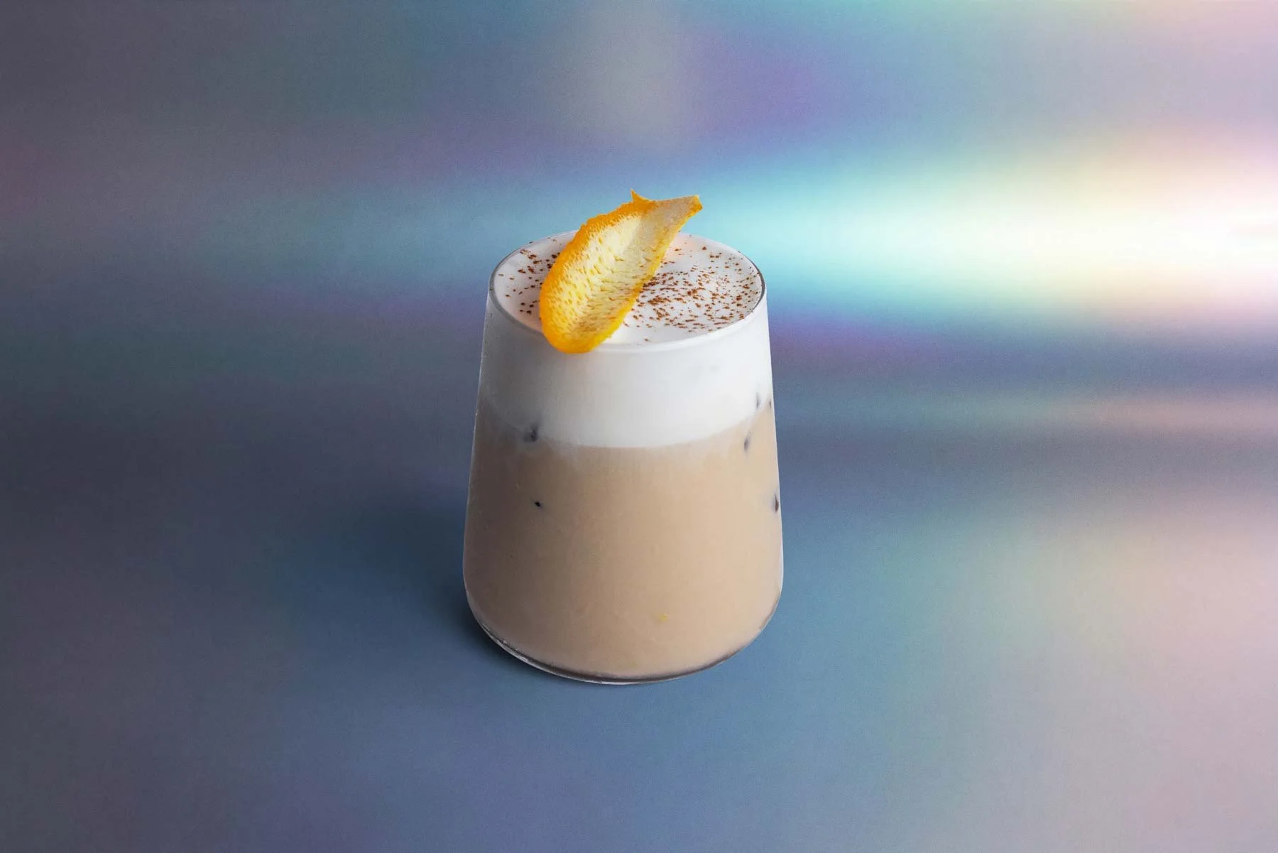 A glass of iced coffee topped with whipped cream, a sprinkle of cocoa powder, and a lemon peel garnish against a gradient colorful background.