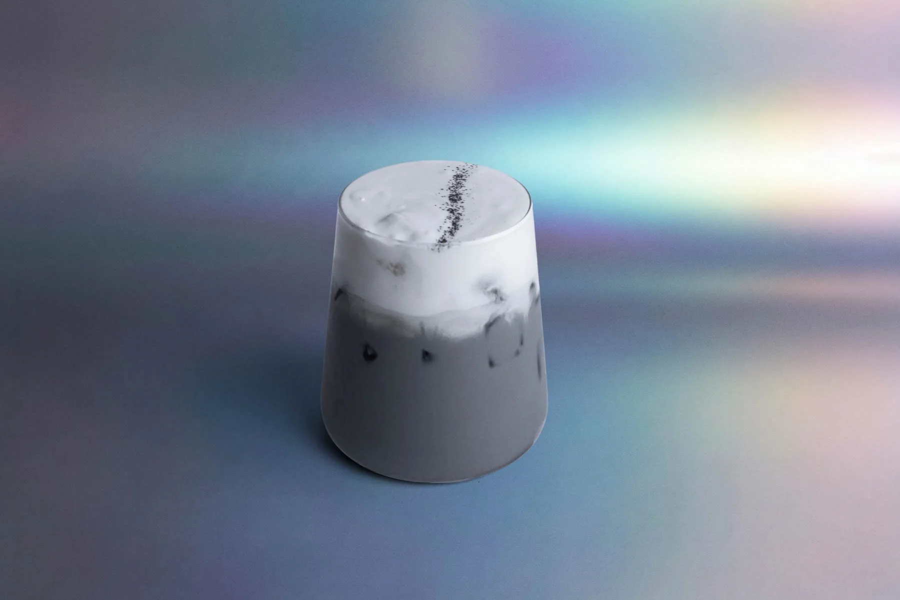 A clear glass with a smoothie topped with whipped cream and a line of black sesame seeds or poppy seeds, set against a colorful, iridescent background.