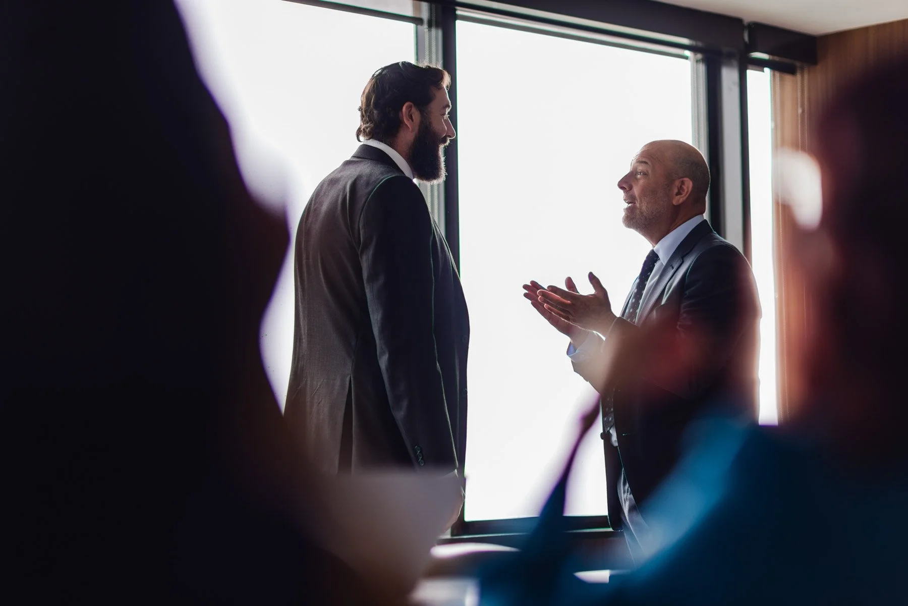 Two men in business suits talking in a bright room with large windows, surrounded by blurred figures.