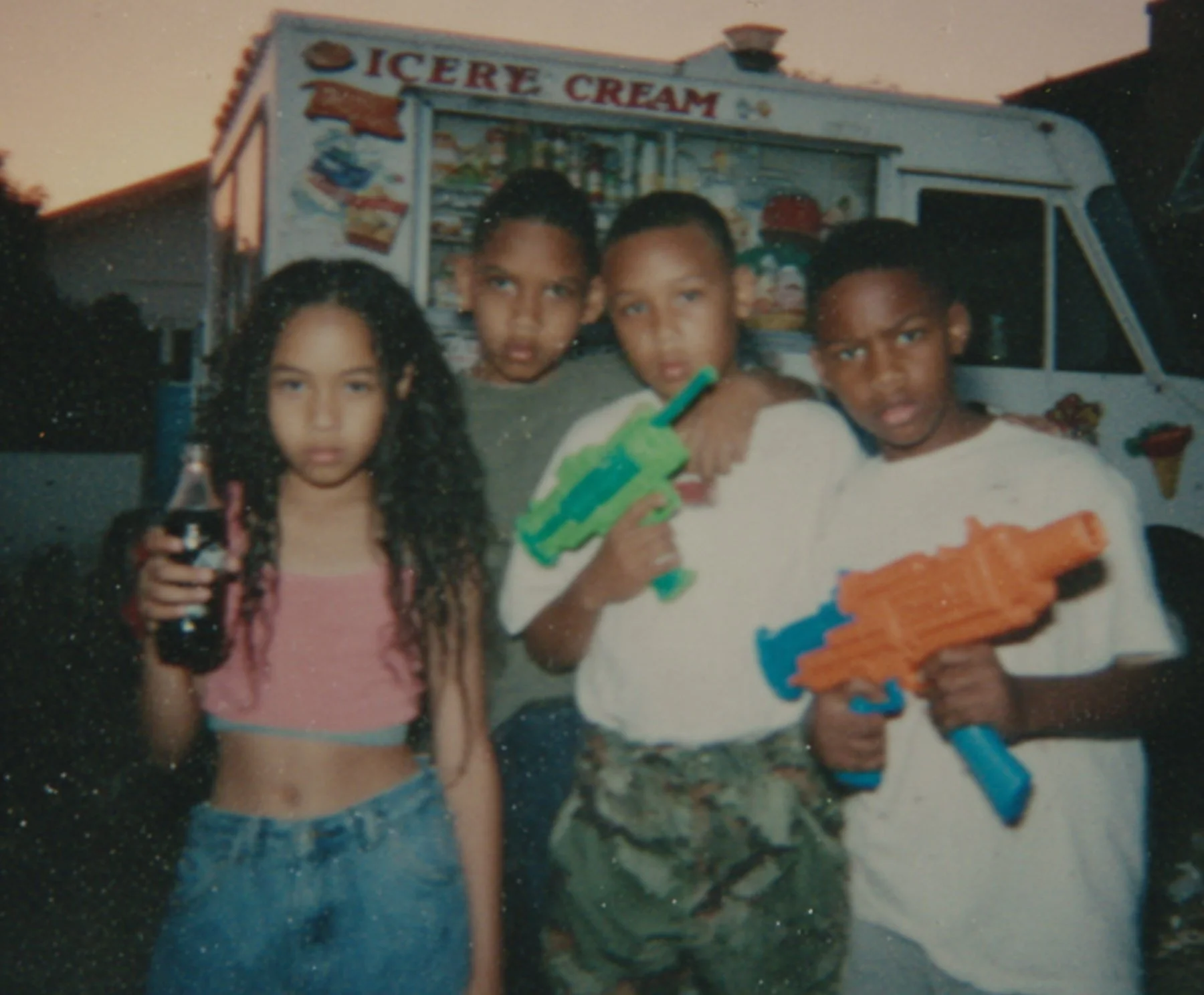 Four children standing in front of an ice cream truck during sunset, holding toy water guns and a beverage, with house rooftops visible in the background.