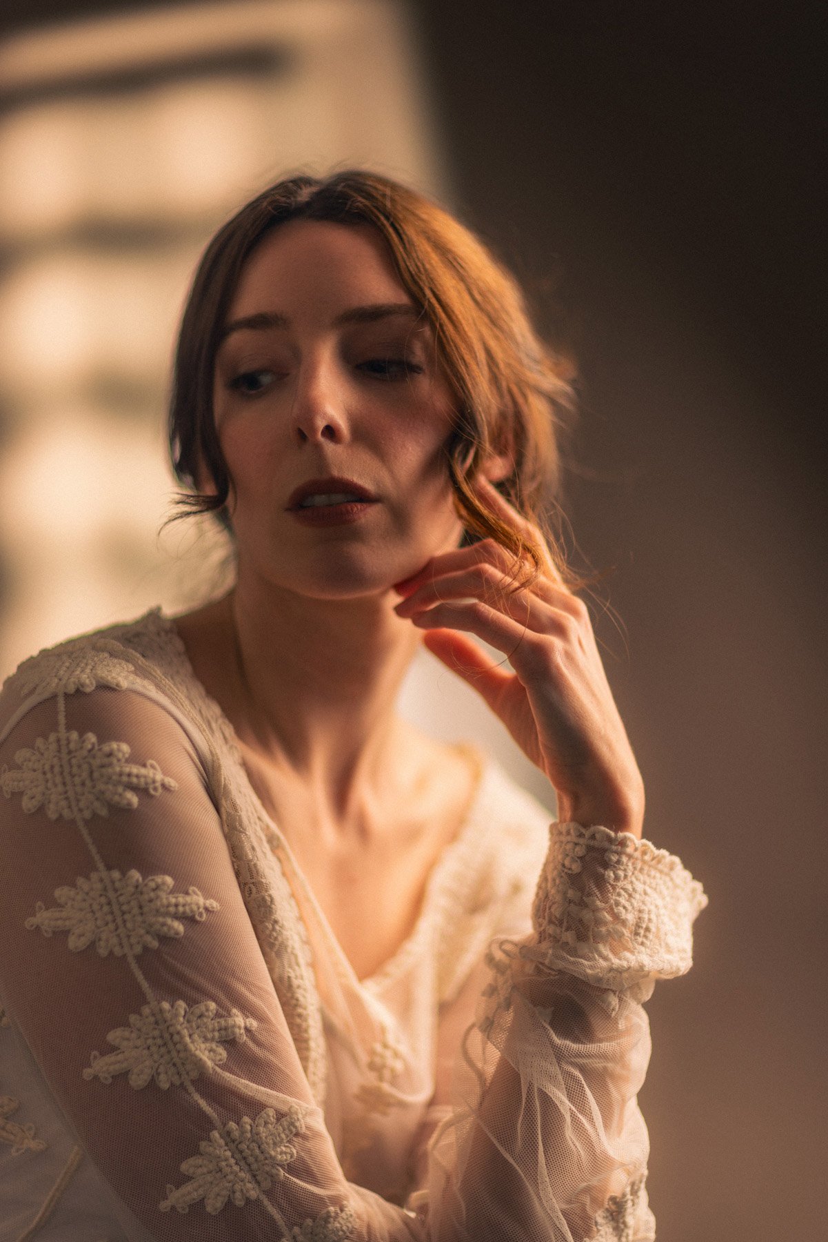 A woman with red hair and fair skin wearing a delicate cream-colored lace dress with floral embroidery, posing indoors with her left hand near her chin, looking thoughtfully to the side.