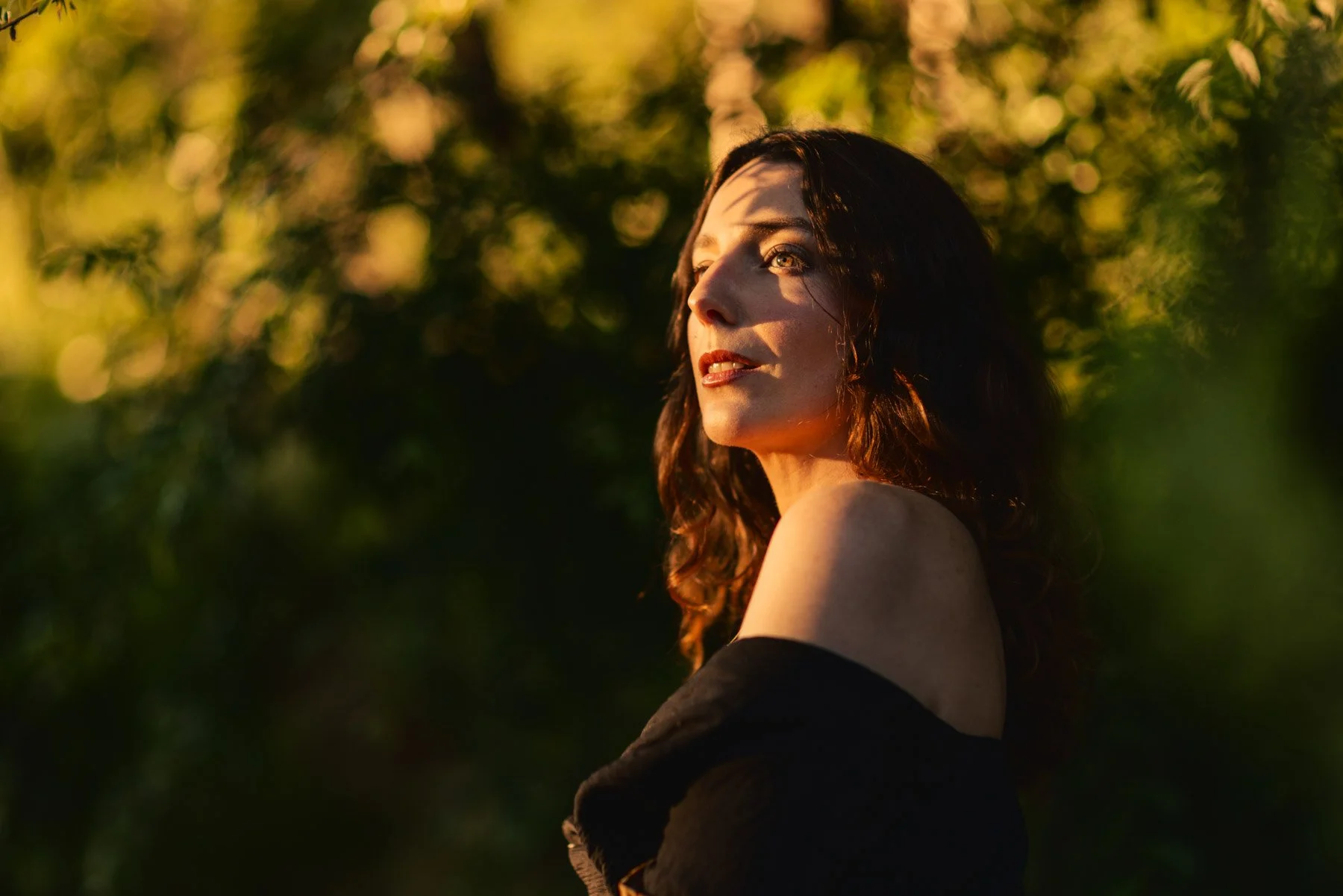 A young woman with wavy dark hair looking to the side with sunlight illuminating her face, standing outdoors with blurred green trees in the background.