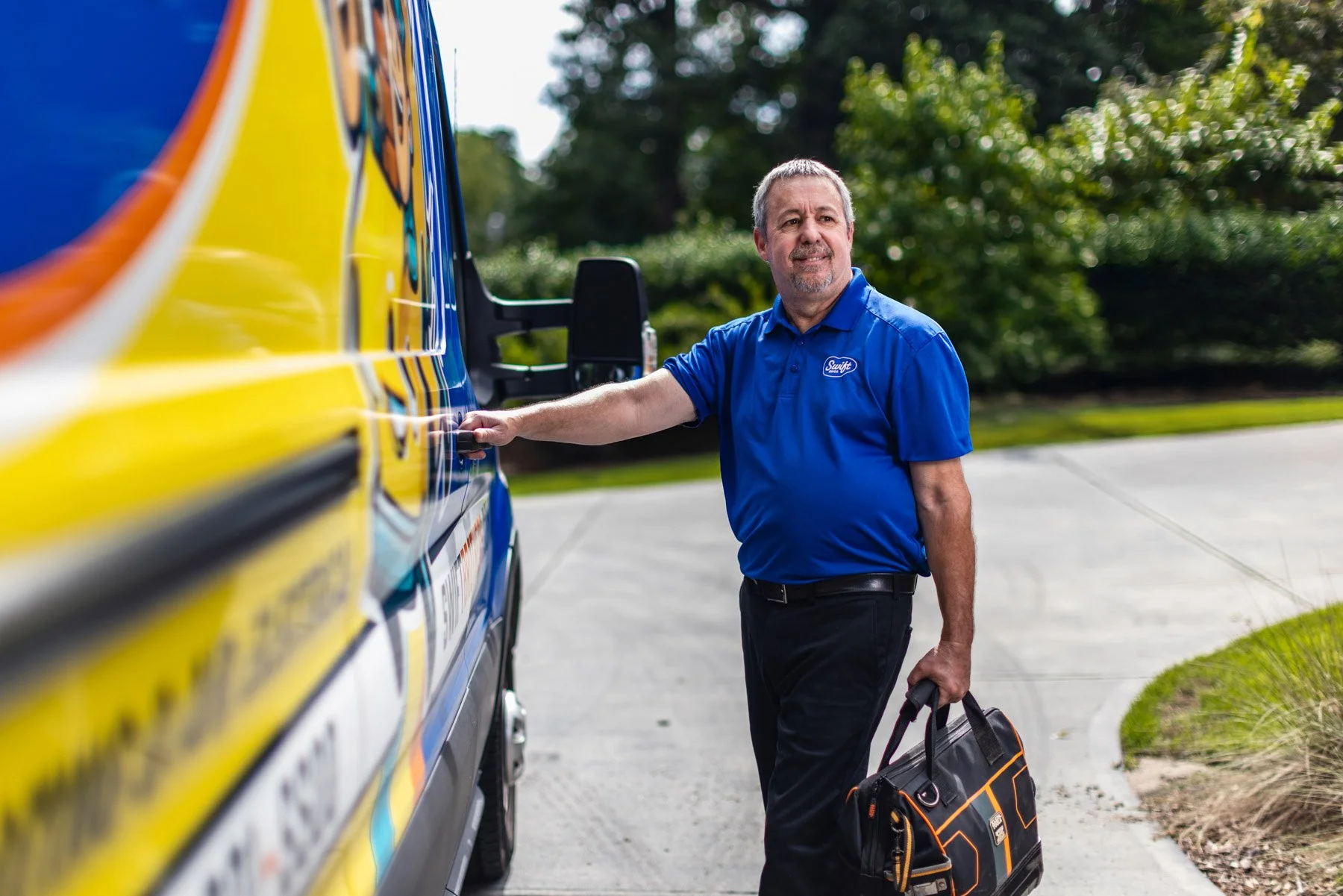 A male technician or delivery worker in a blue uniform pressing a button on a yellow appliance truck, holding a black and orange bag, in an outdoor parking area with trees in the background.