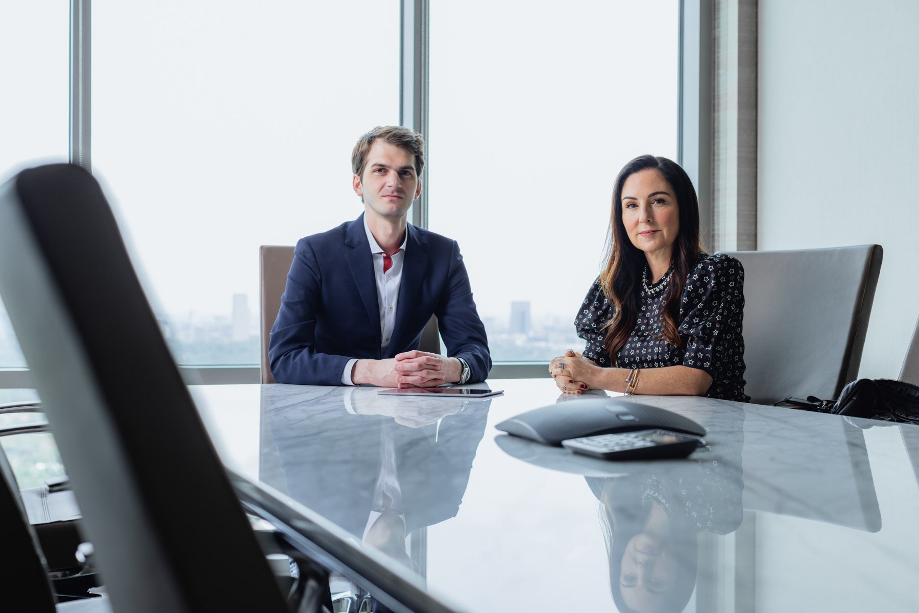 A man and a woman seated at a conference table in a modern office with large windows, a city skyline view, a speakerphone, and a tablet in front of them.