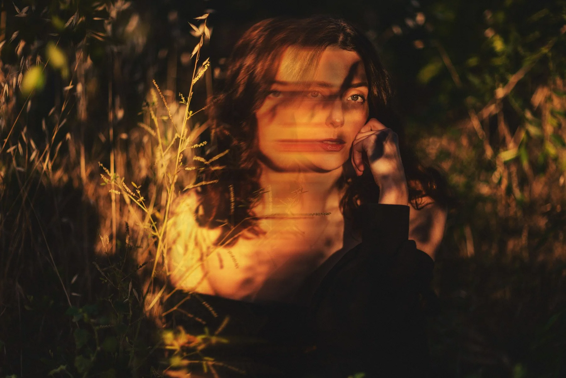 A woman with dark curly hair and fair skin is sitting amidst tall grasses and plants at dusk, illuminated by a warm, golden light that creates a dreamy effect. She rests her chin on her hand and looks softly at the camera.