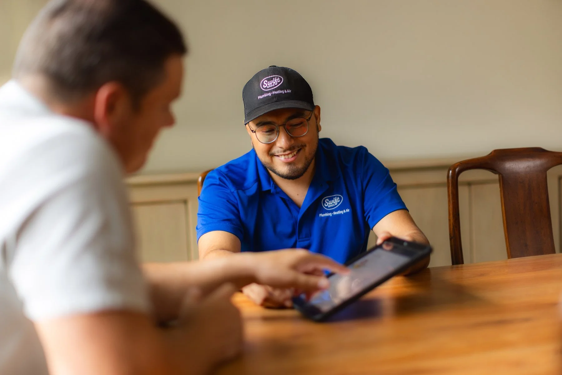 A smiling man in a blue Swift Bros technician uniform and cap showing a phone to another person during a consultation at a wooden table in an office setting.