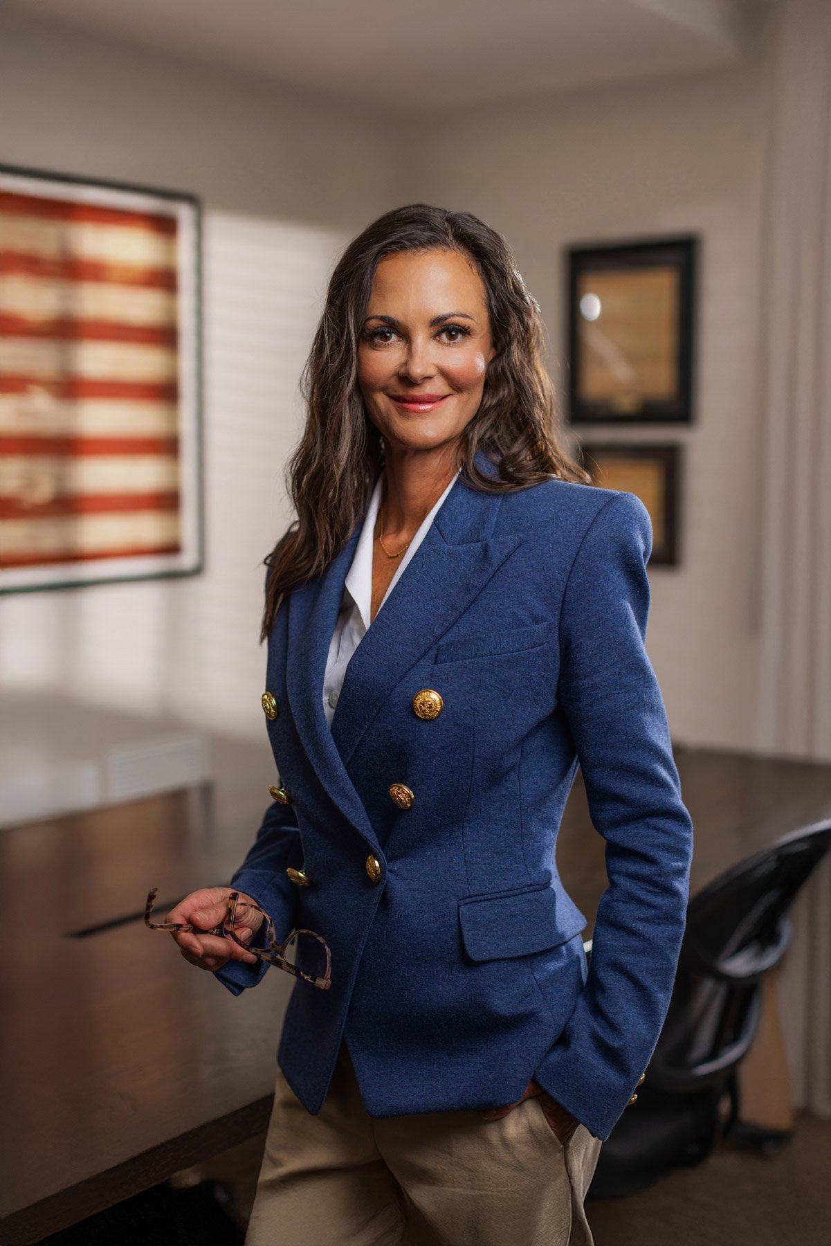 A woman with wavy hair wearing a navy blue blazer with gold buttons, holding eyeglasses, standing indoors with artwork on the wall behind her.