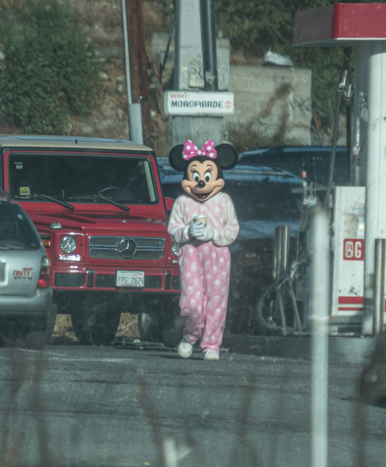 Someone wearing a Minnie Mouse costume in pink pajamas with white polka dots, holding a coffee cup, standing in front of a row of parked cars at a gas station.