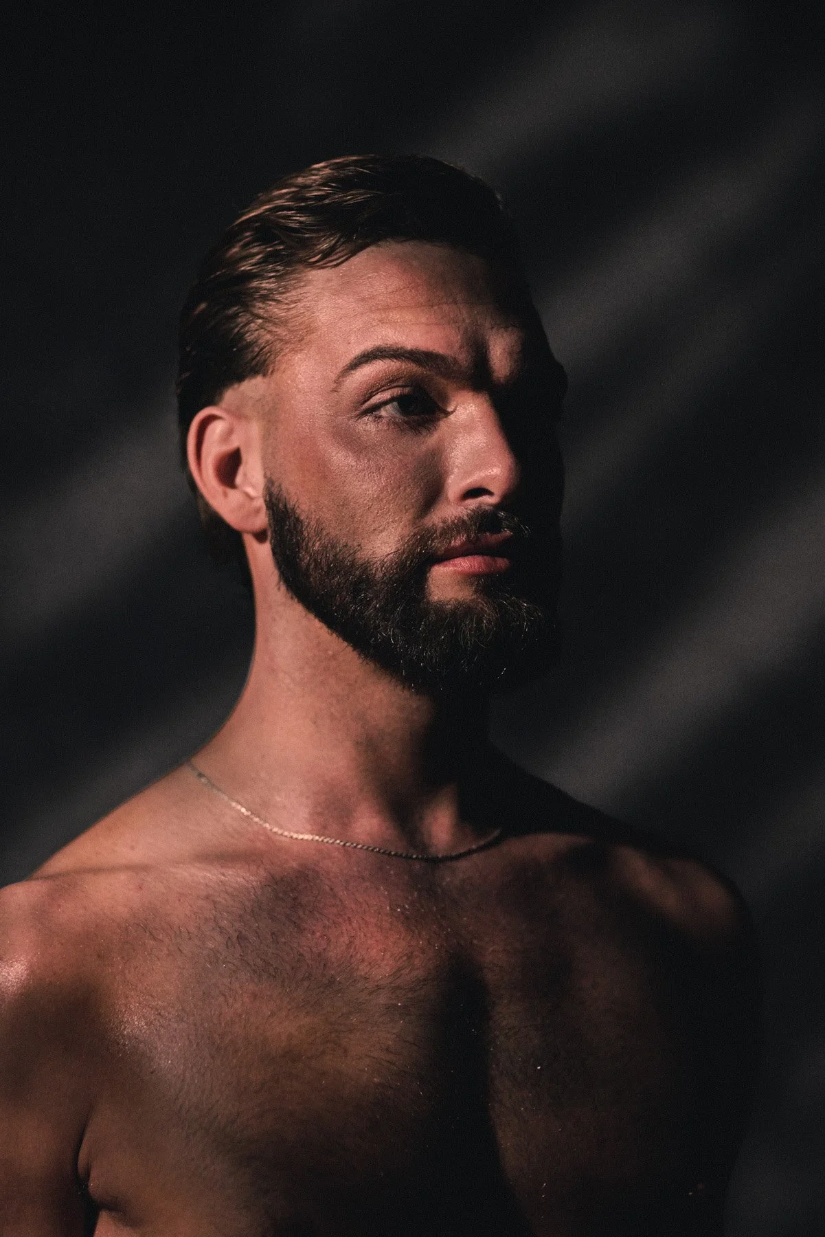 A shirtless man with a beard and mustache, looking to the side, with dramatic lighting creating shadows on his face and upper body.