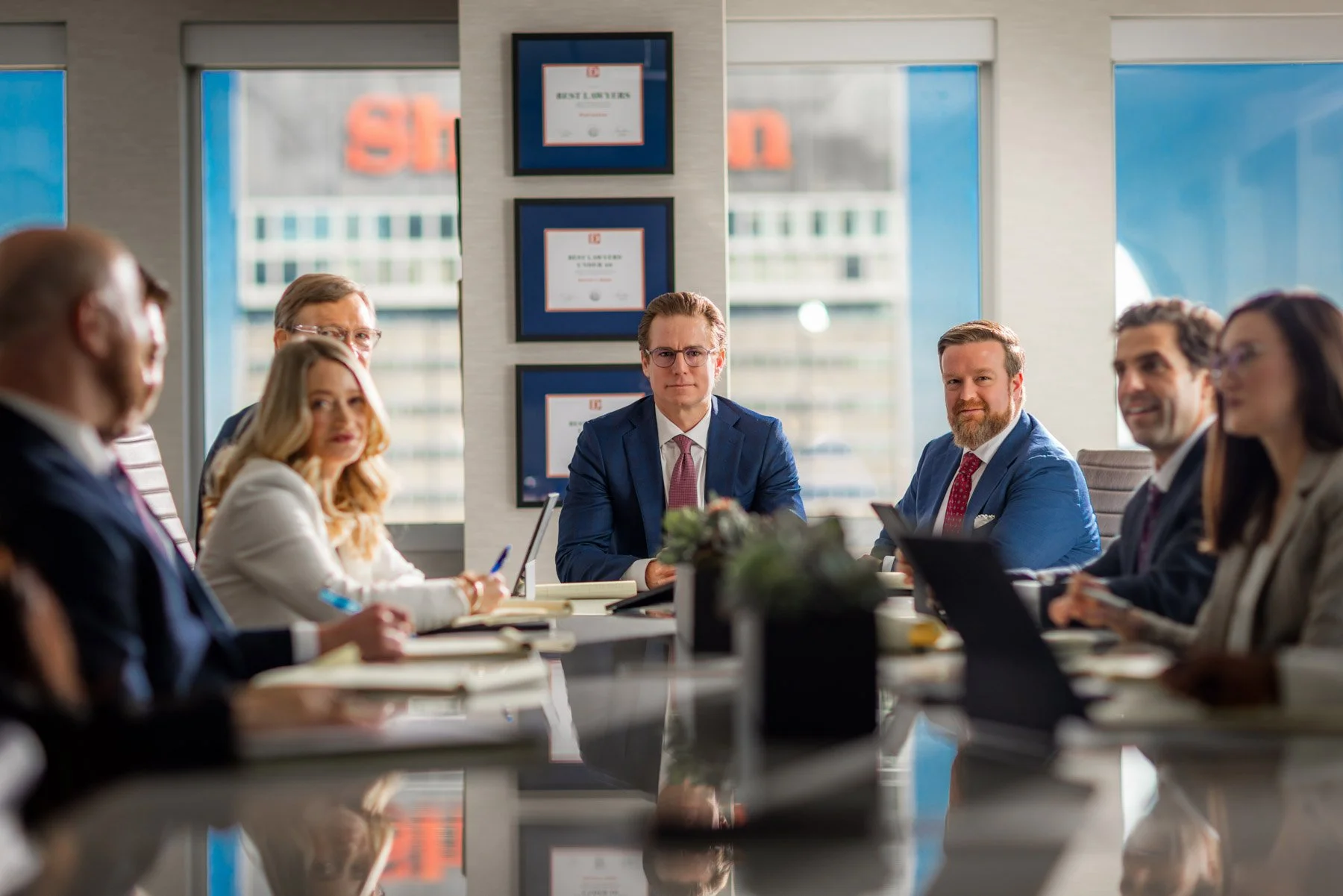 Business meeting in a modern conference room with seven people seated around a table. A man in a suit and glasses sit at the head of the table, with other professionals listening and engaging in discussion. Large windows let in natural light, and fra