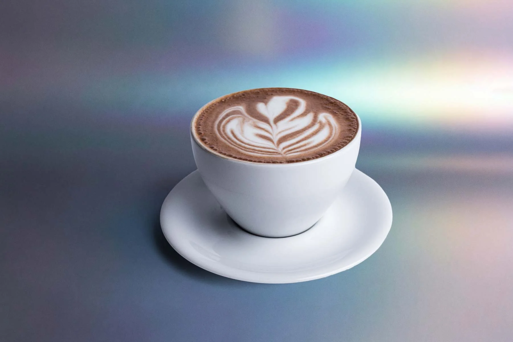 A white cup of hot chocolate with latte art in the shape of a heart and floral design, resting on a matching white saucer on a smooth, reflective surface.