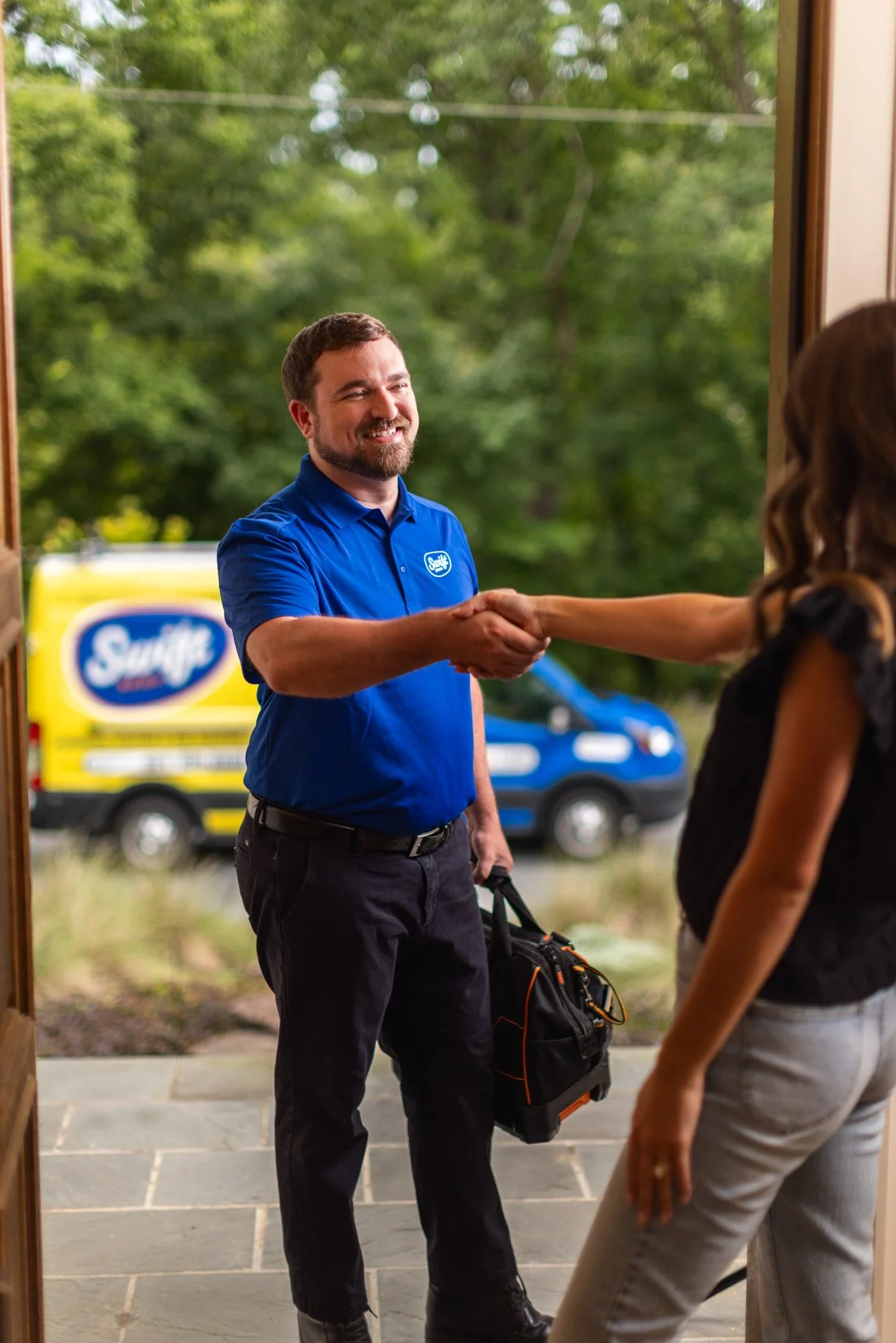 A male delivery worker with a beard wearing a blue uniform shirt shaking hands with a woman inside a home. The delivery person is holding a black and orange bag. In the background, there is a yellow and blue Swift delivery truck and a blue vehicle pa
