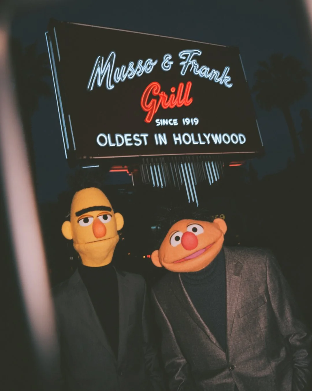 Two people wearing puppets of Bert and Ernie from Sesame Street, standing in front of a neon sign for Musso & Frank Grill, a historic restaurant in Hollywood.