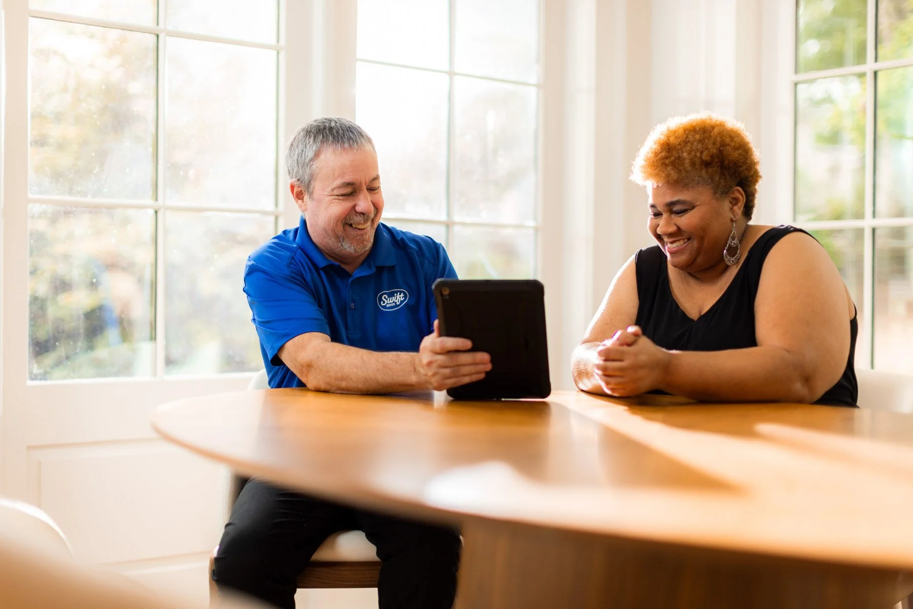 A smiling man in a blue shirt showing something on a tablet to a woman smiling and looking at the tablet, sitting at a table near a window.