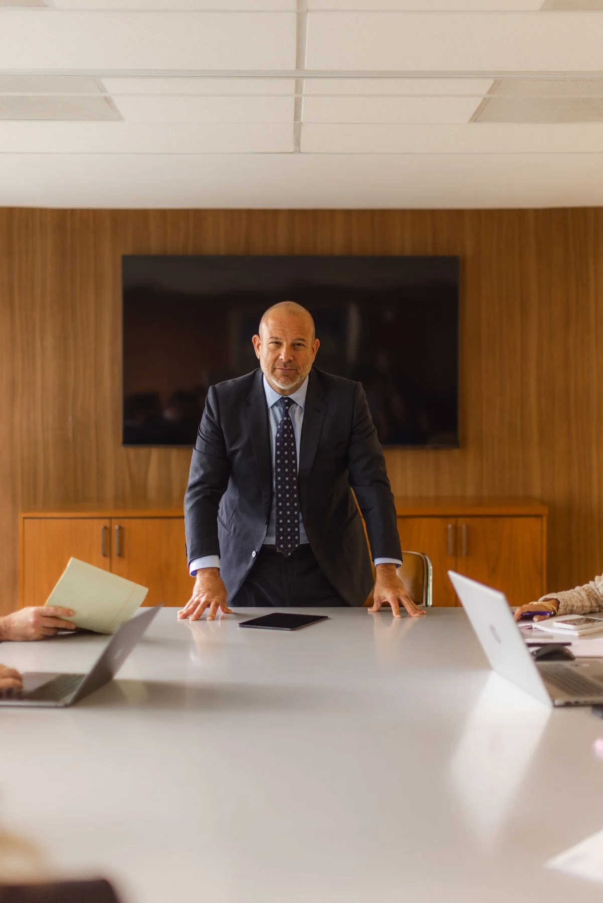 A man in a business suit standing in a conference room behind a table with laptops and documents, with a large TV screen behind him.