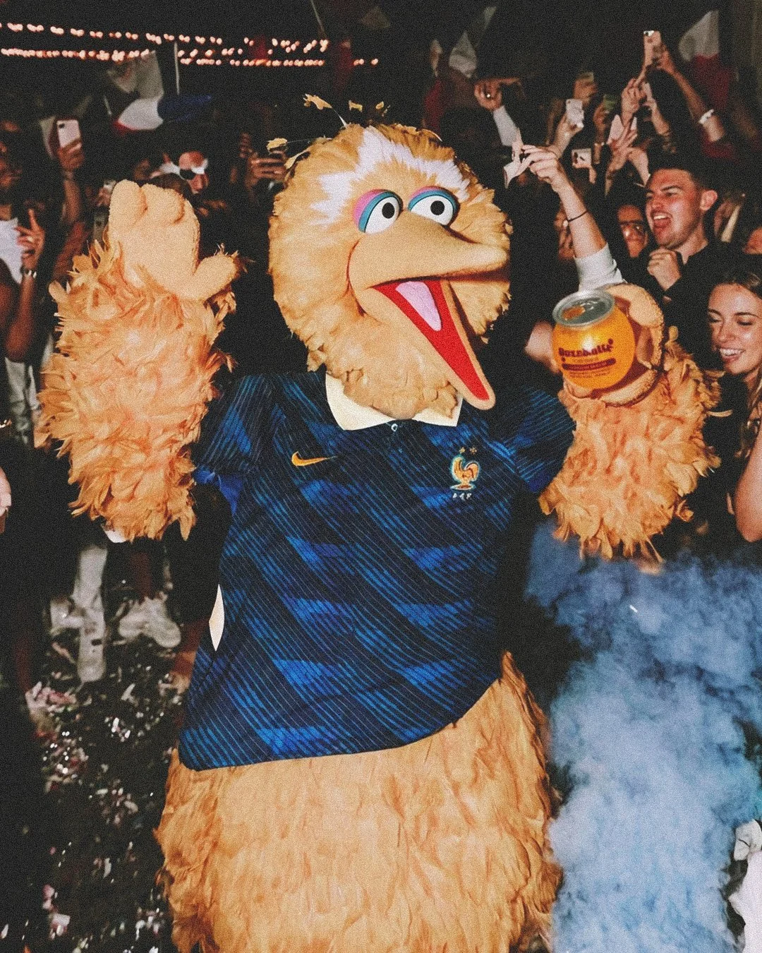 Sesame Streets Big Bird, wearing a blue French national soccer team jersey, holding a buzzball, surrounded by cheering crowd at a lively event.
