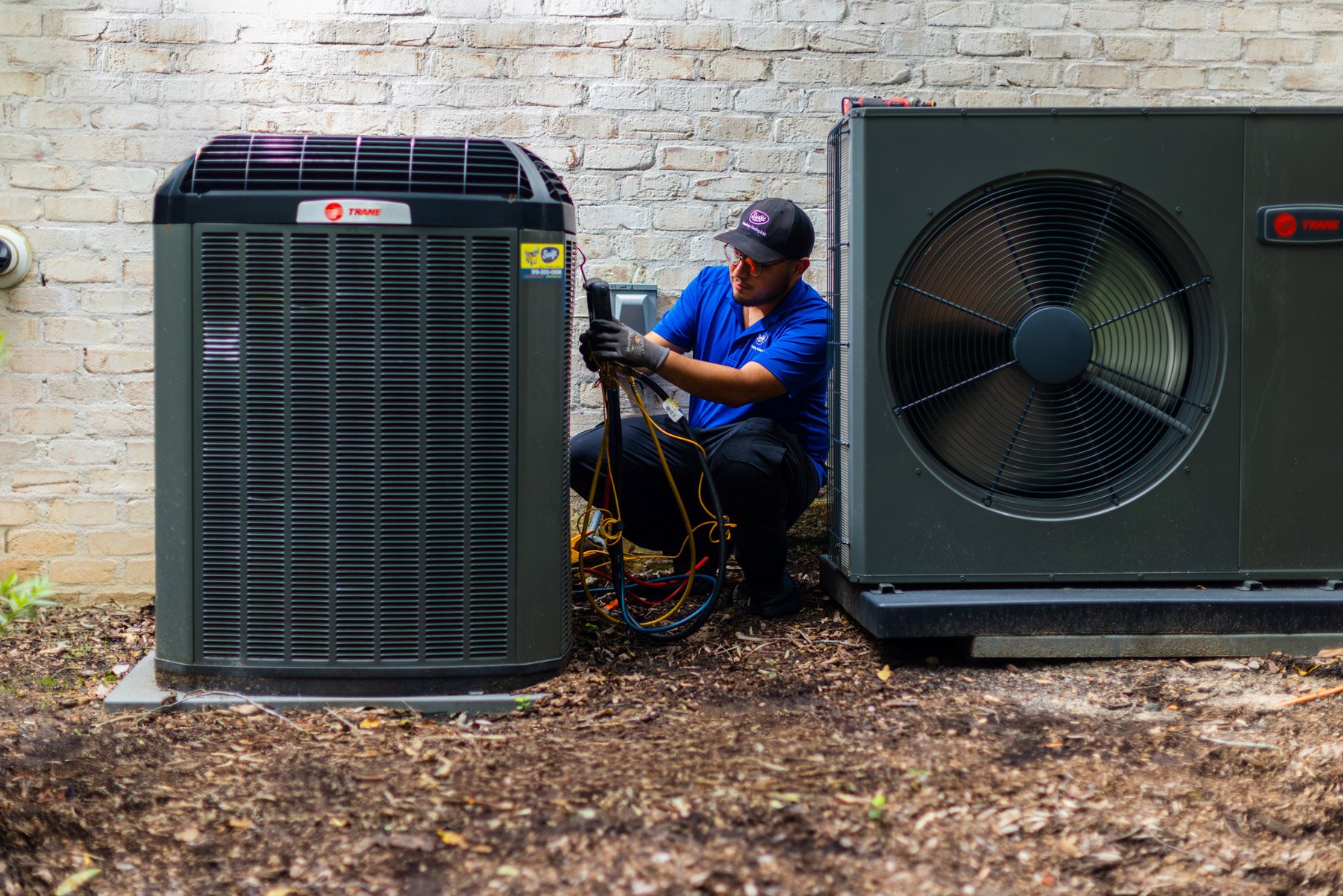 Technician working on an outdoor HVAC unit, kneeling with tools and wires near a brick wall.