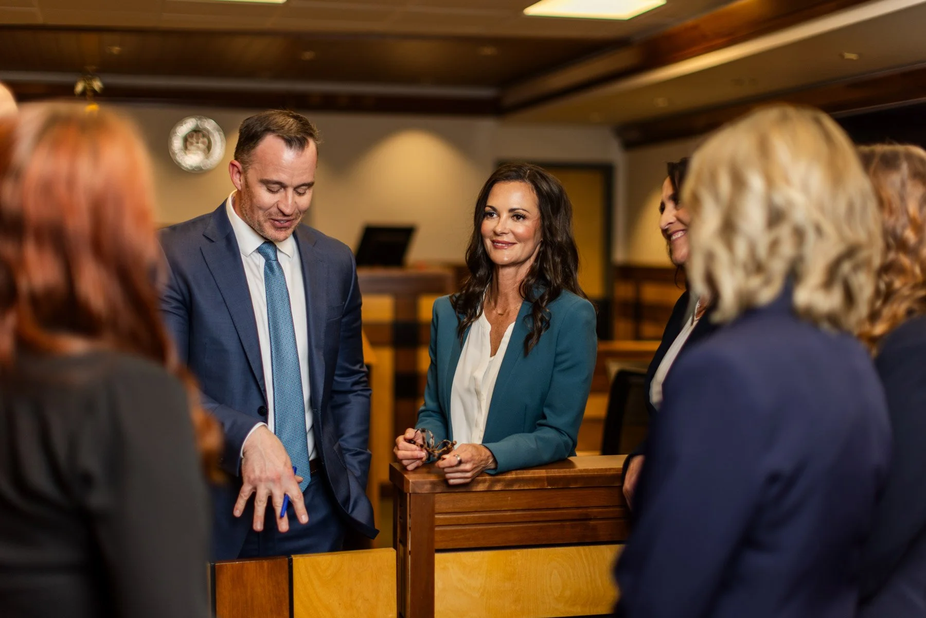 Group of people in a formal setting, engaged in conversation; some women are wearing dark blazers, and a man in a blue suit is showing something on his hand.
